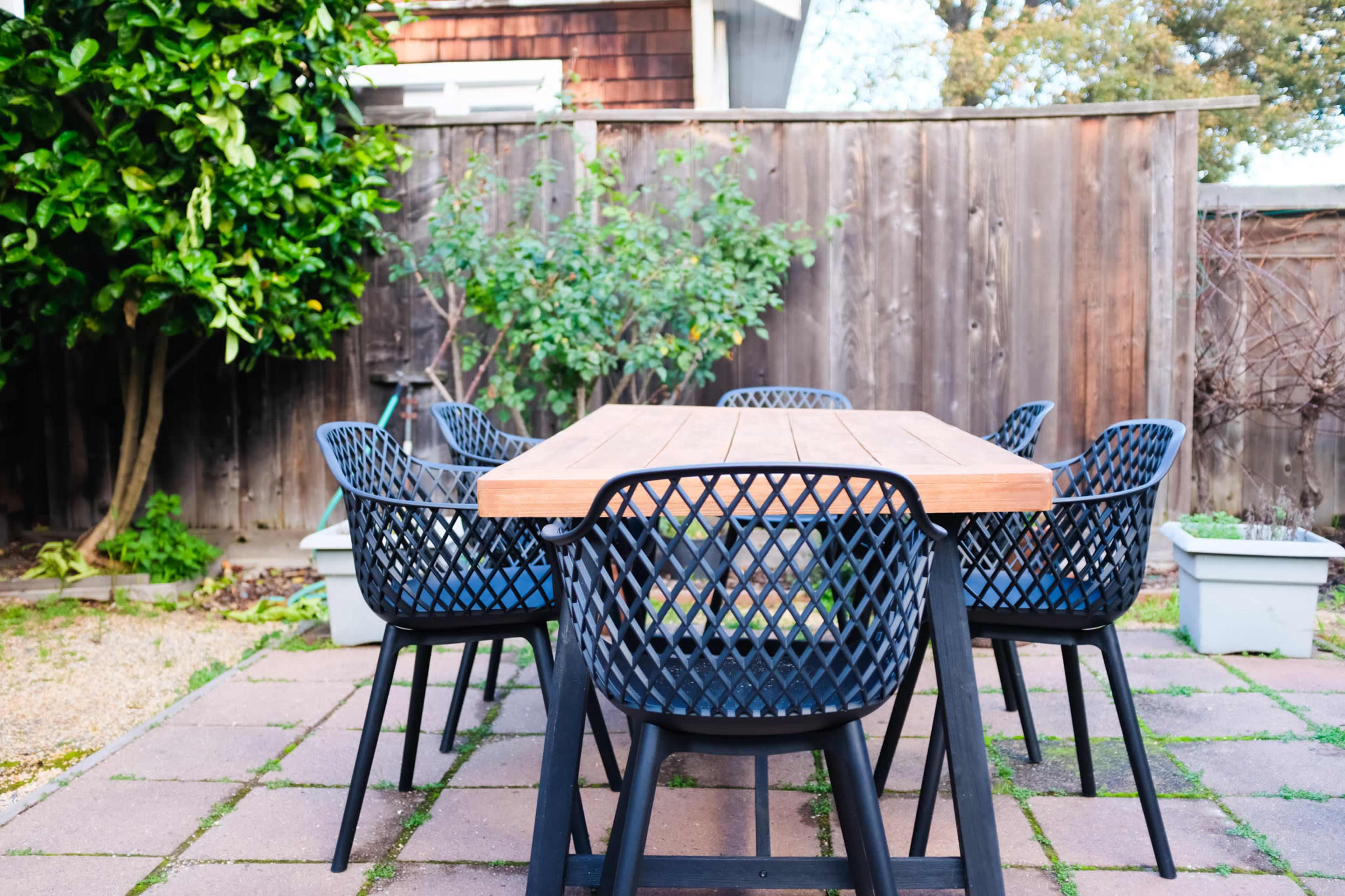 A wooden table surrounded by six black woven chairs is set on a paved patio, with greenery and a wooden fence in the background.