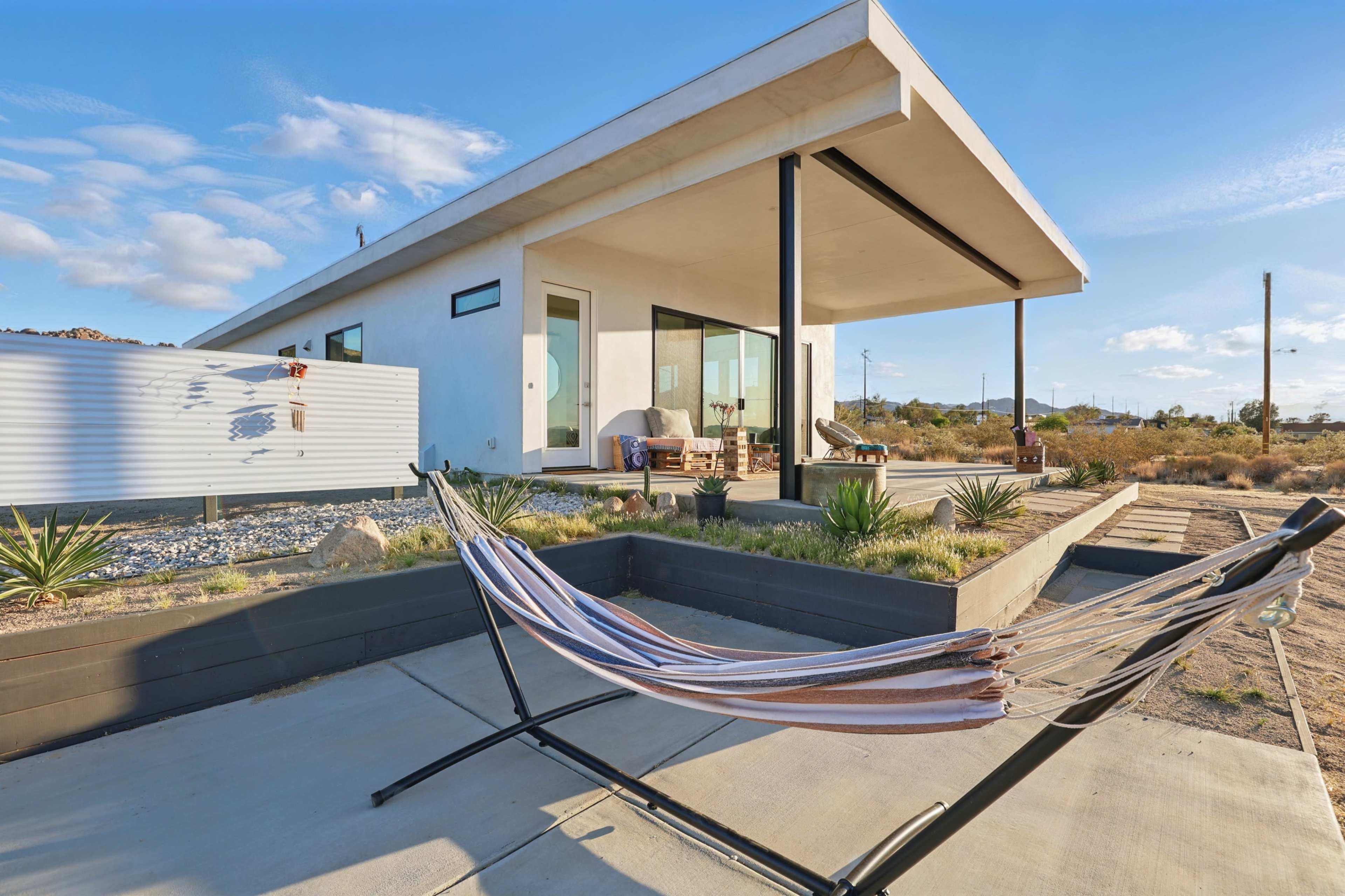 A modern house with large windows and a flat roof is situated in a desert landscape, featuring a hammock set in front of a landscaped area.