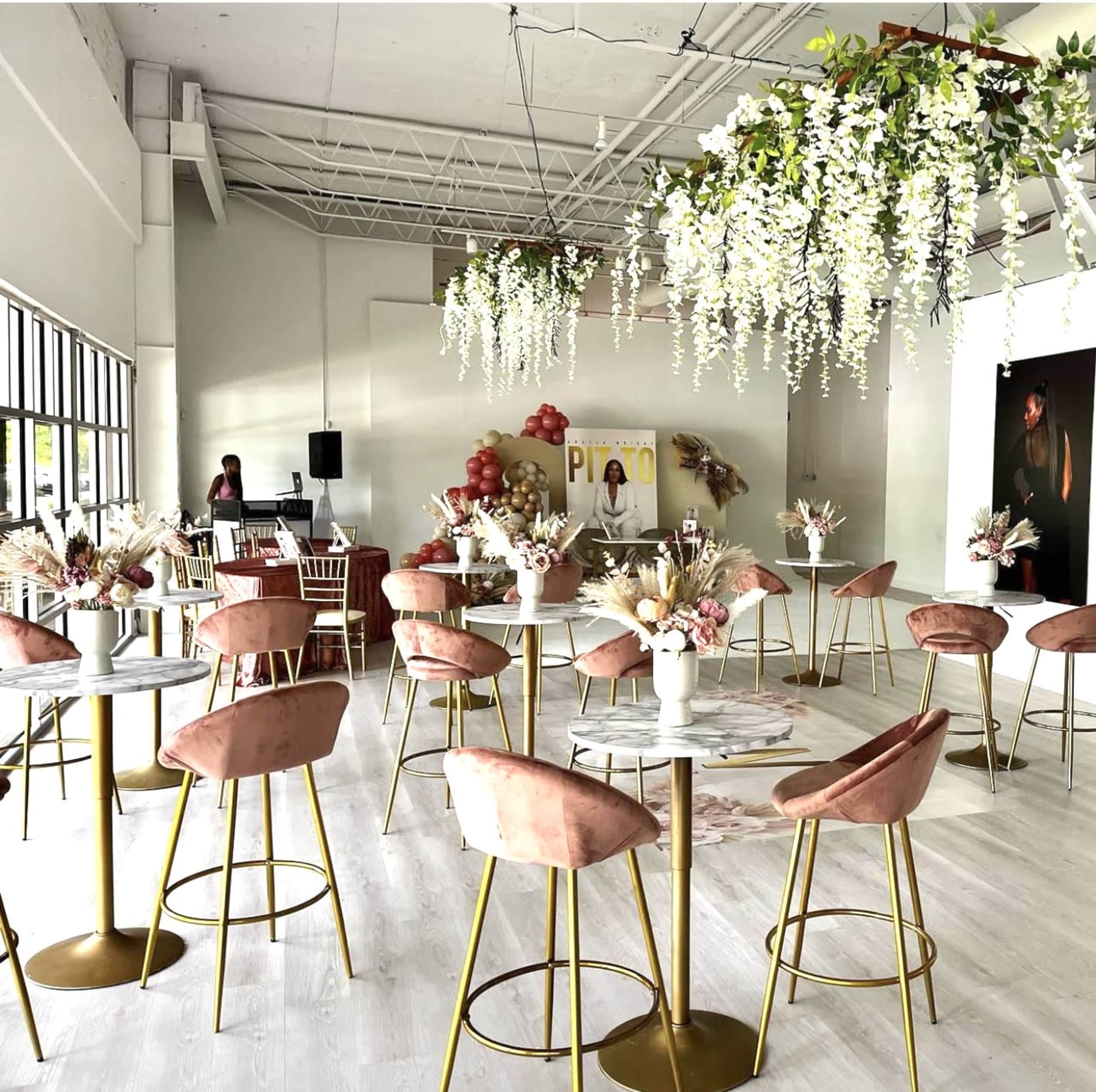 The image shows a modern event space with round tables featuring pink upholstered chairs, floral centerpieces, and hanging greenery decorations.