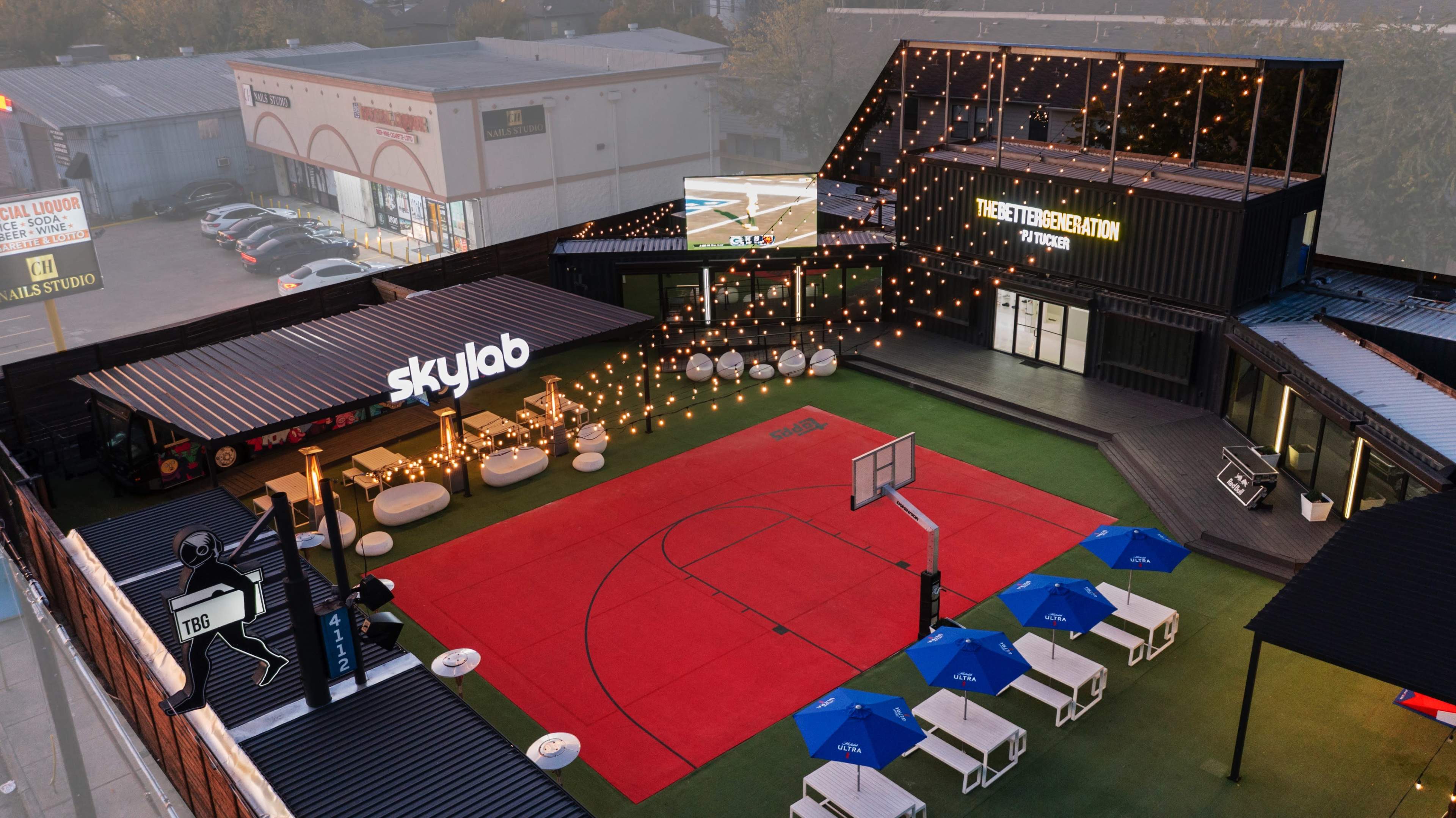 The scene shows an outdoor recreational area featuring a red basketball court, white seating, and blue umbrellas, surrounded by a modern building with bright signage and string lights.