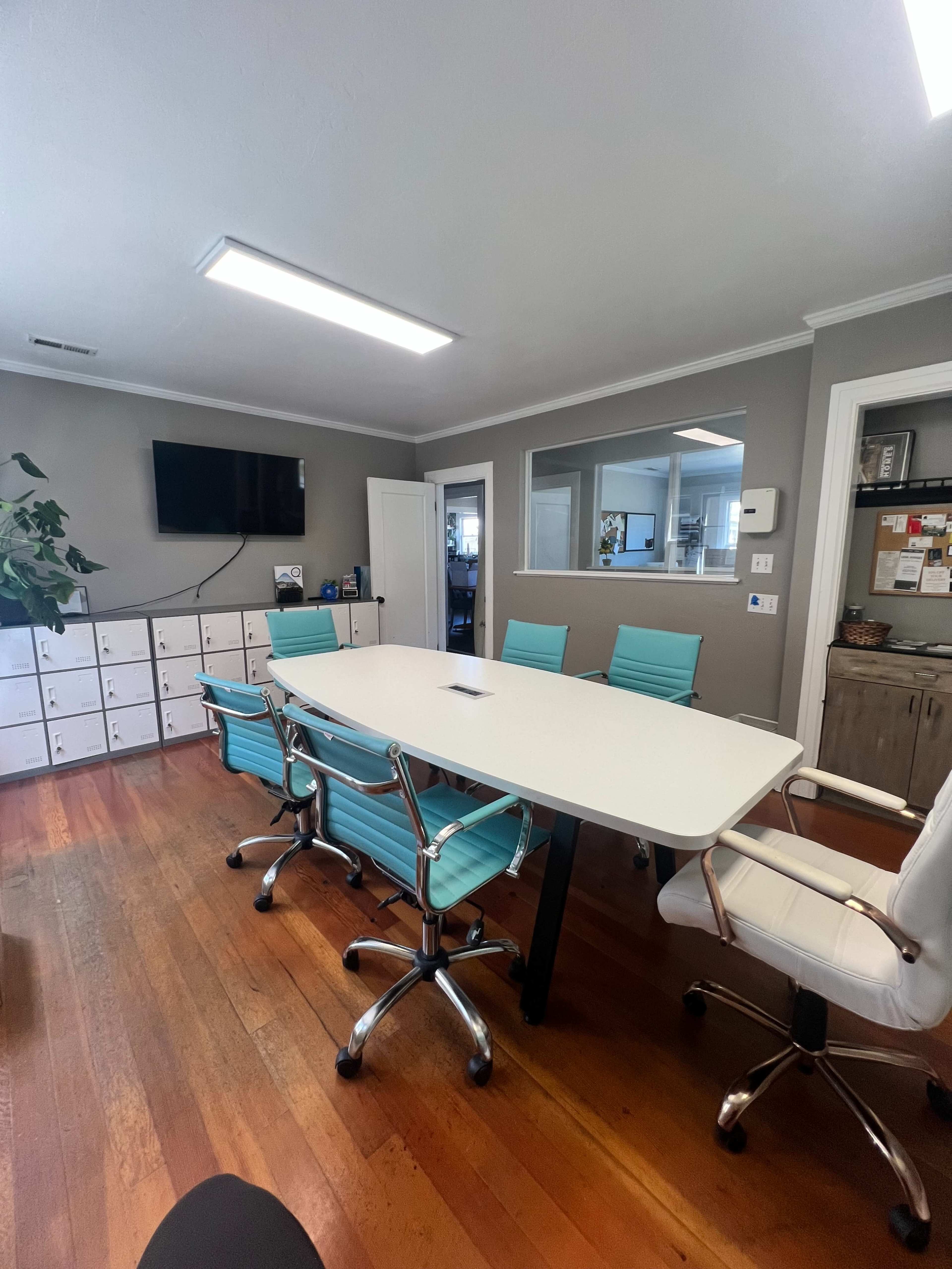 A modern conference room features a white table surrounded by turquoise chairs, with a large TV mounted on the wall and a cabinet of drawers in the background.