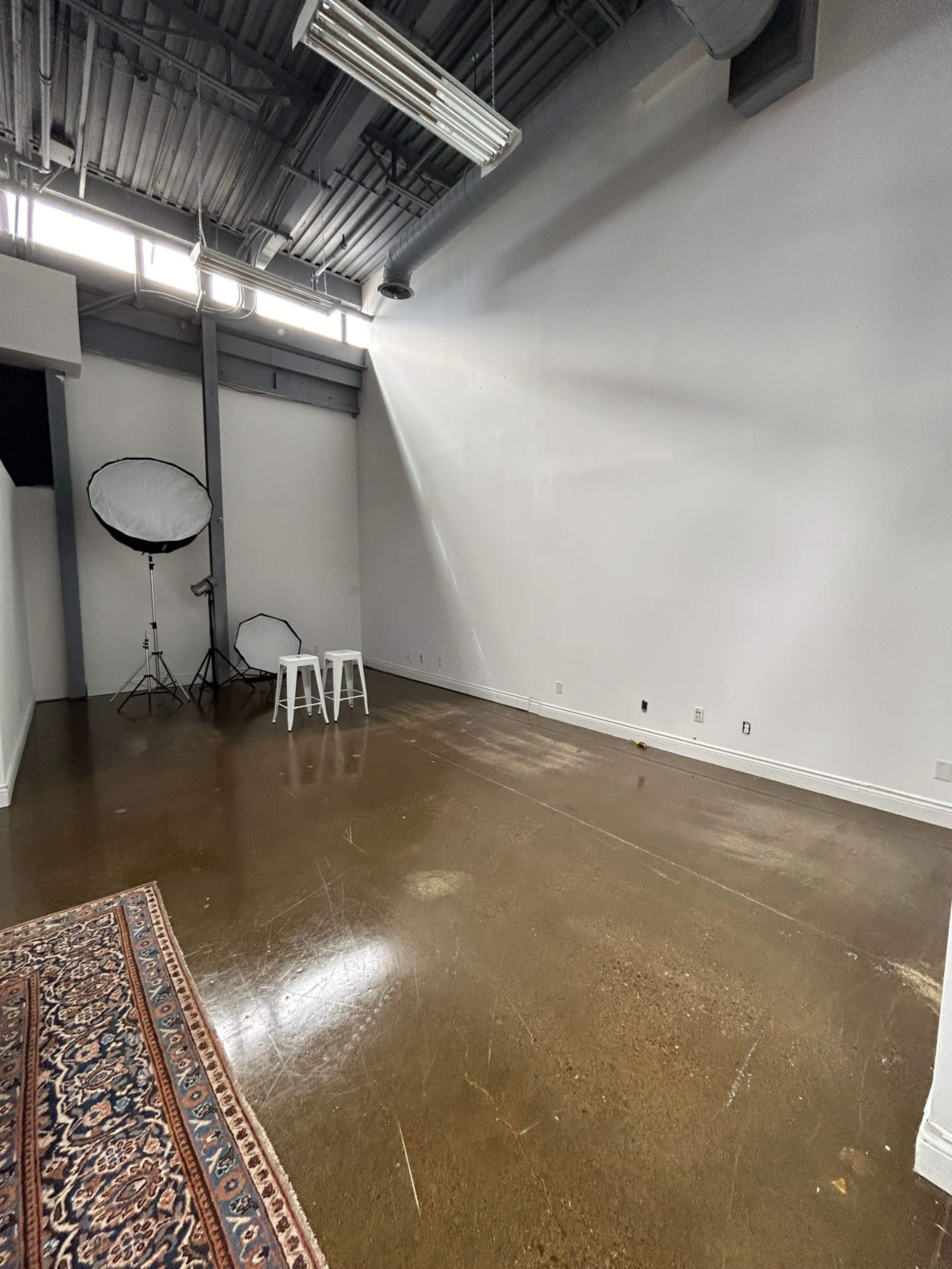The room features a polished concrete floor, two white stools, and a large lighting setup in the corner, against a plain white wall.