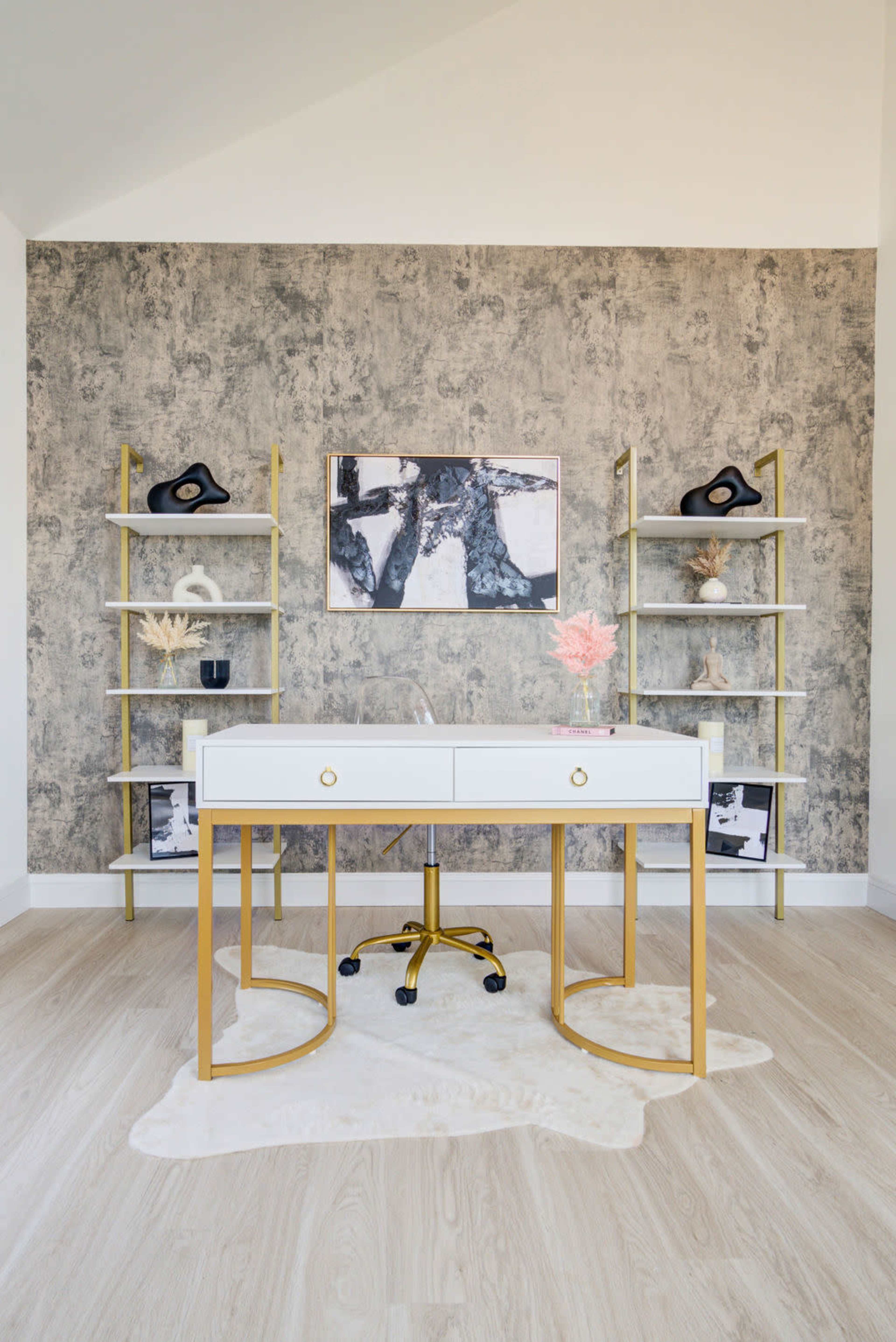 A modern office space features a white desk with gold accents, surrounded by two vertical shelves holding decorative items, against a textured gray wall.
