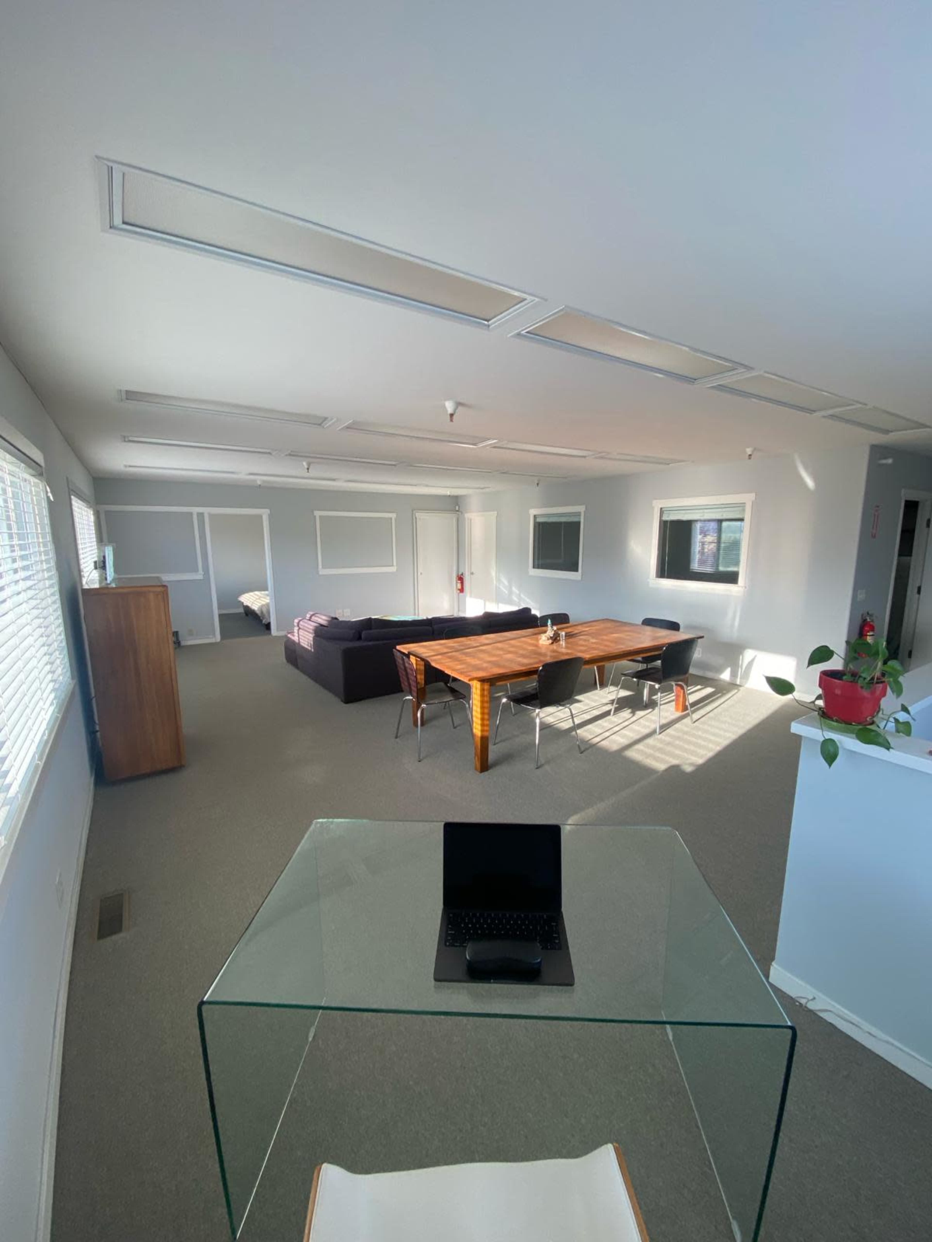 The image shows a spacious office interior with a dining table and chairs, a seating area, and large windows allowing natural light.