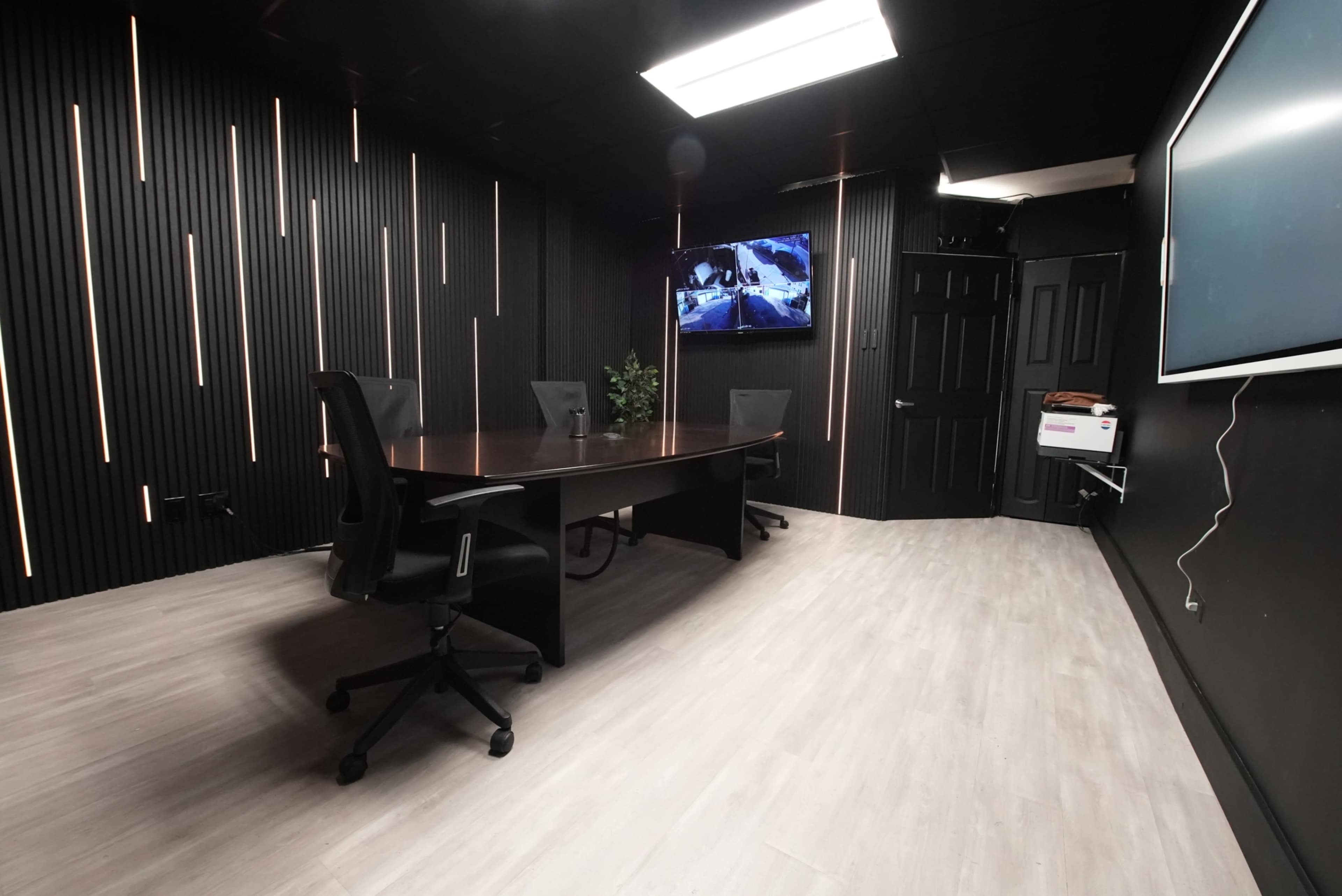 The image shows a modern conference room with a long table, black panelled walls, and four office chairs positioned around the table.