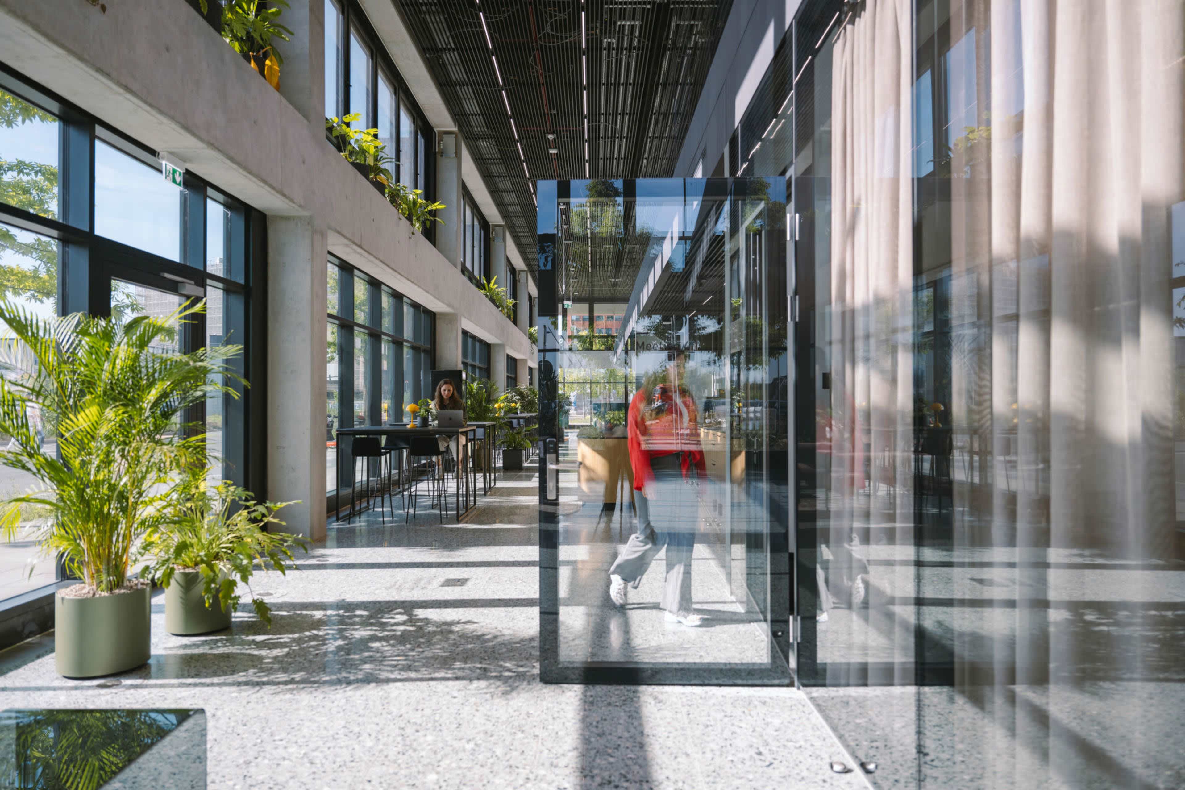 The image shows a bright, modern interior space with large windows, plants, and a glass entrance.