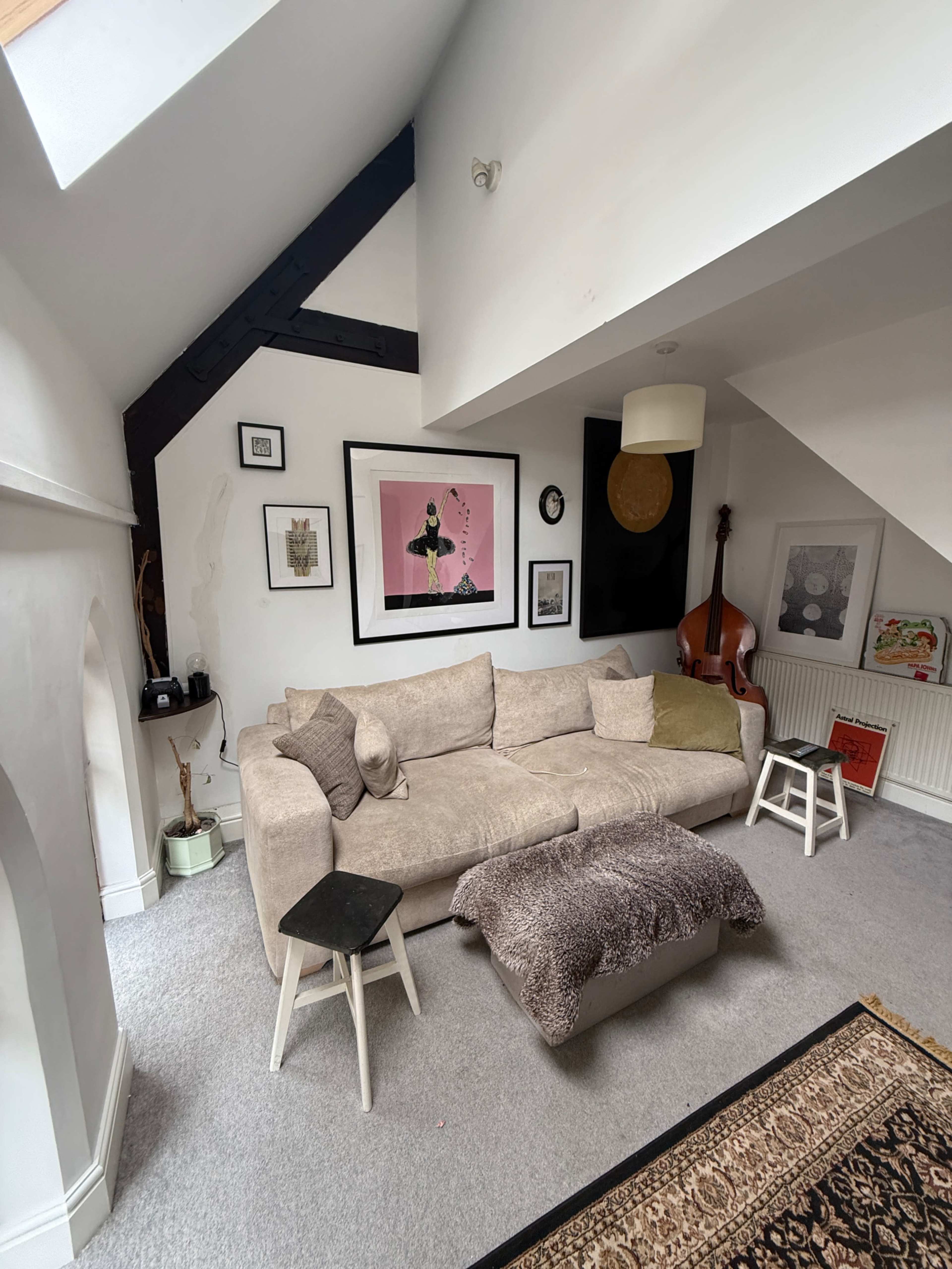 The image shows a cozy living room with a beige sectional sofa, various framed artworks on the walls, and a corner featuring a double bass.