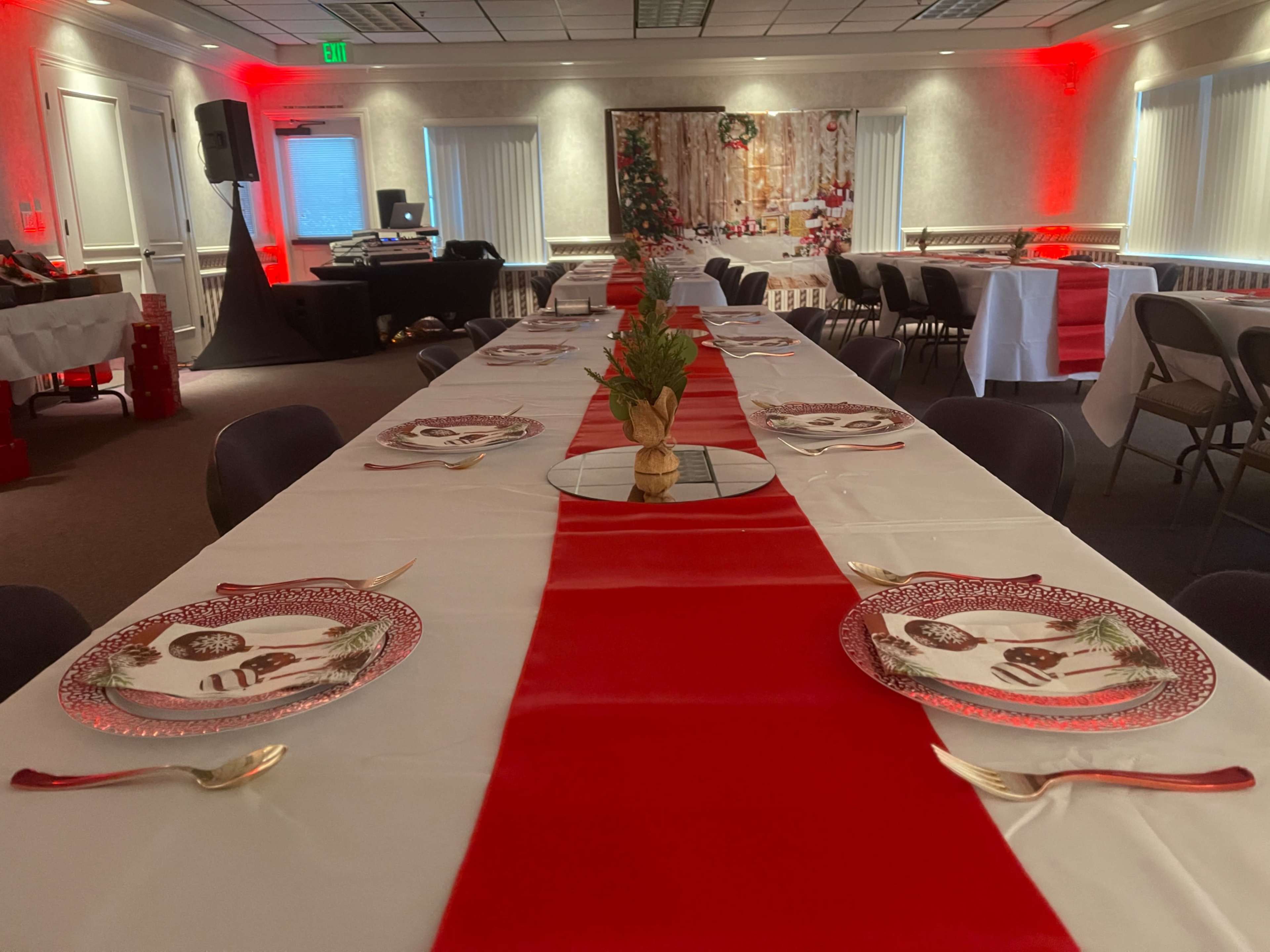 A long dining table is set with holiday-themed plates and decorated with a red table runner in a festively lit room.