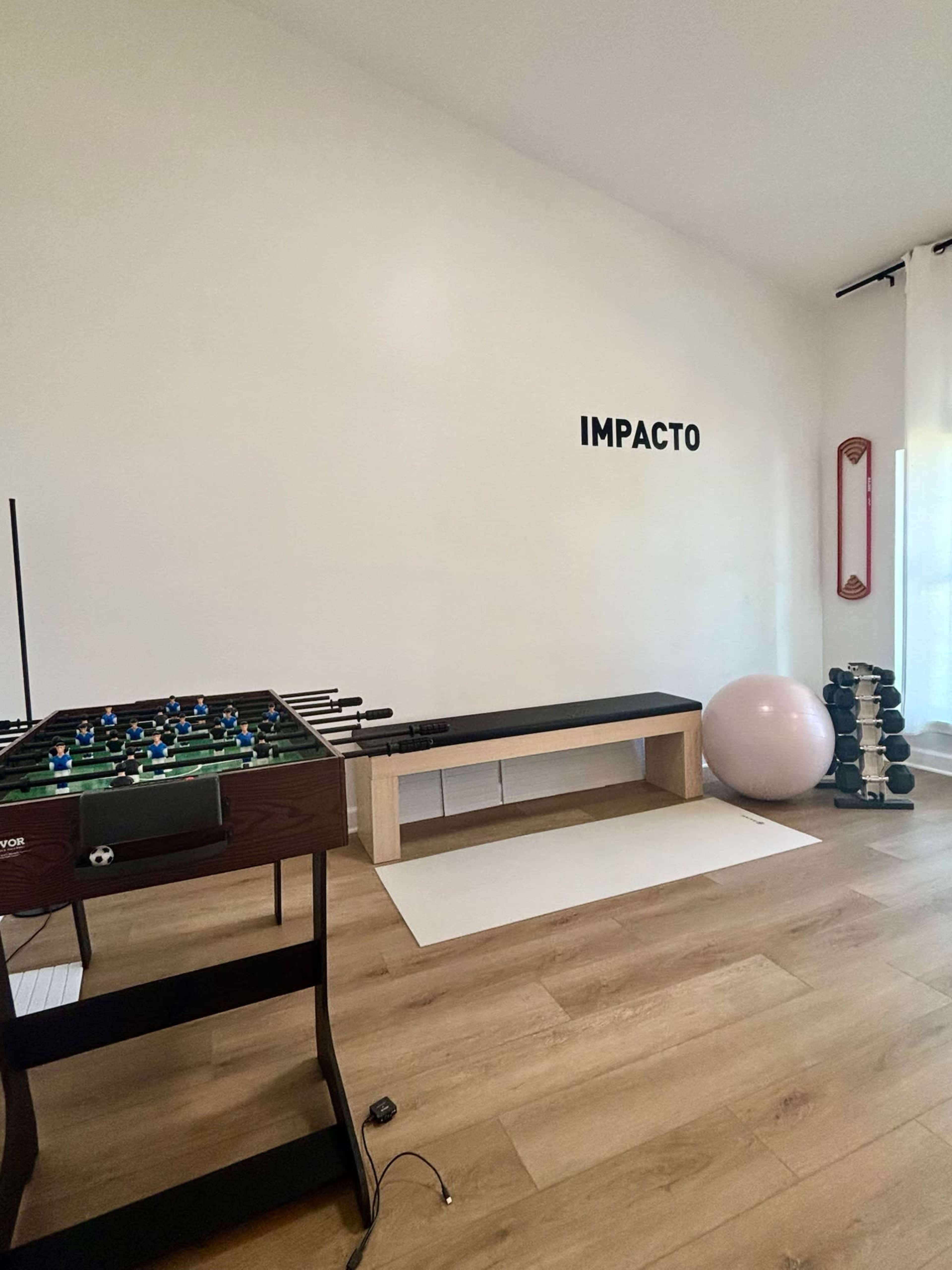 The image shows a room with a foosball table, a bench, an exercise ball, dumbbells, and the word "IMPACTO" displayed on the wall.