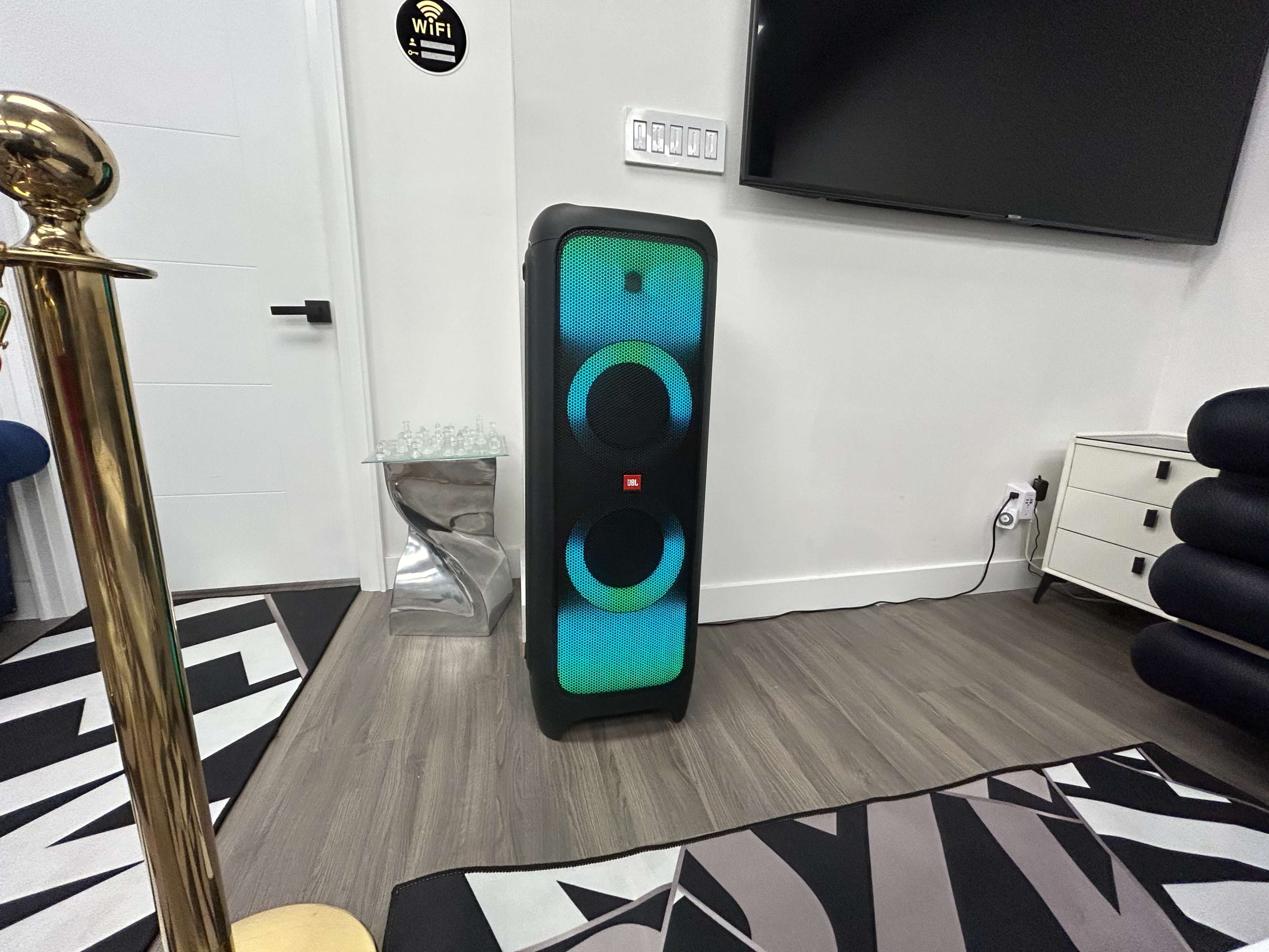 A large black speaker with colorful LED lights stands on a hardwood floor next to a modern TV in a stylish interior.