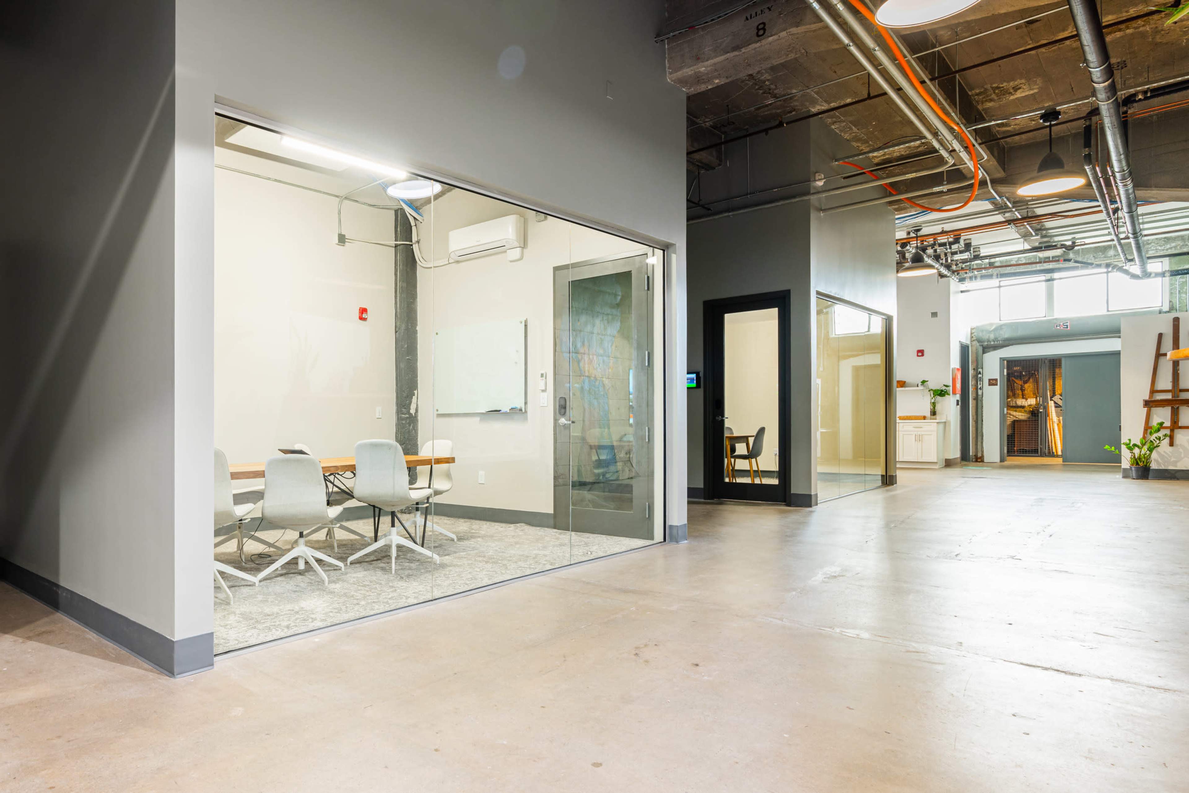 The image shows a modern interior space with a glass-walled conference room on the left and an office door on the right, all situated within a large open area.