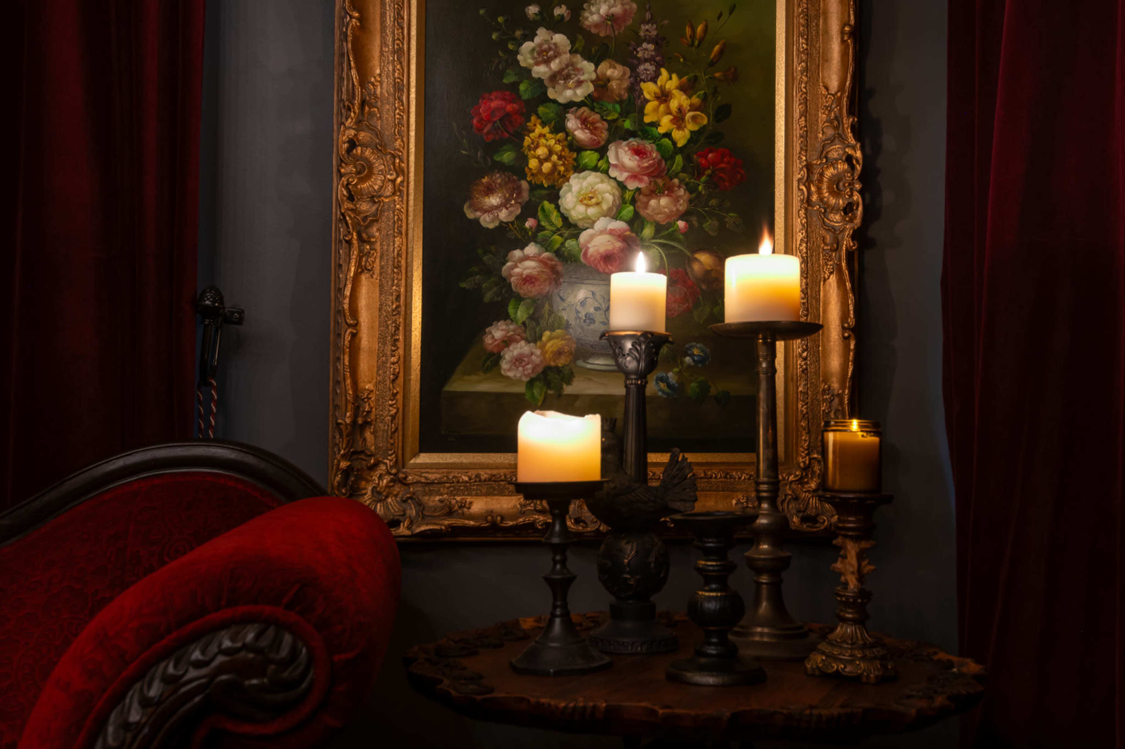 A red upholstered chair is positioned beside a wooden table adorned with various candle holders and lit candles, set against a backdrop featuring a floral painting framed in ornate gold.