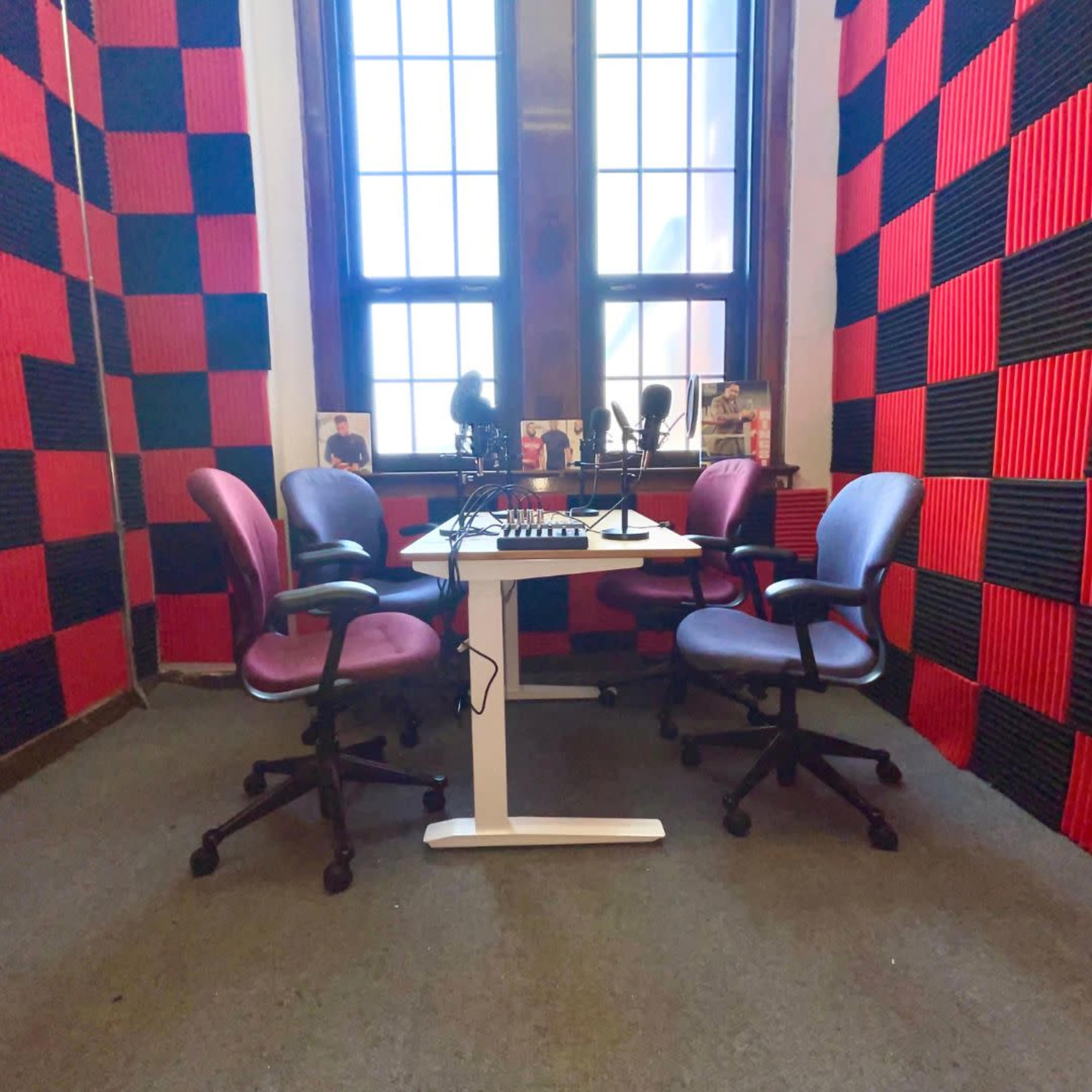A recording studio with a white table and four rolling chairs, surrounded by red and black acoustic foam panels, and two microphones on the table.