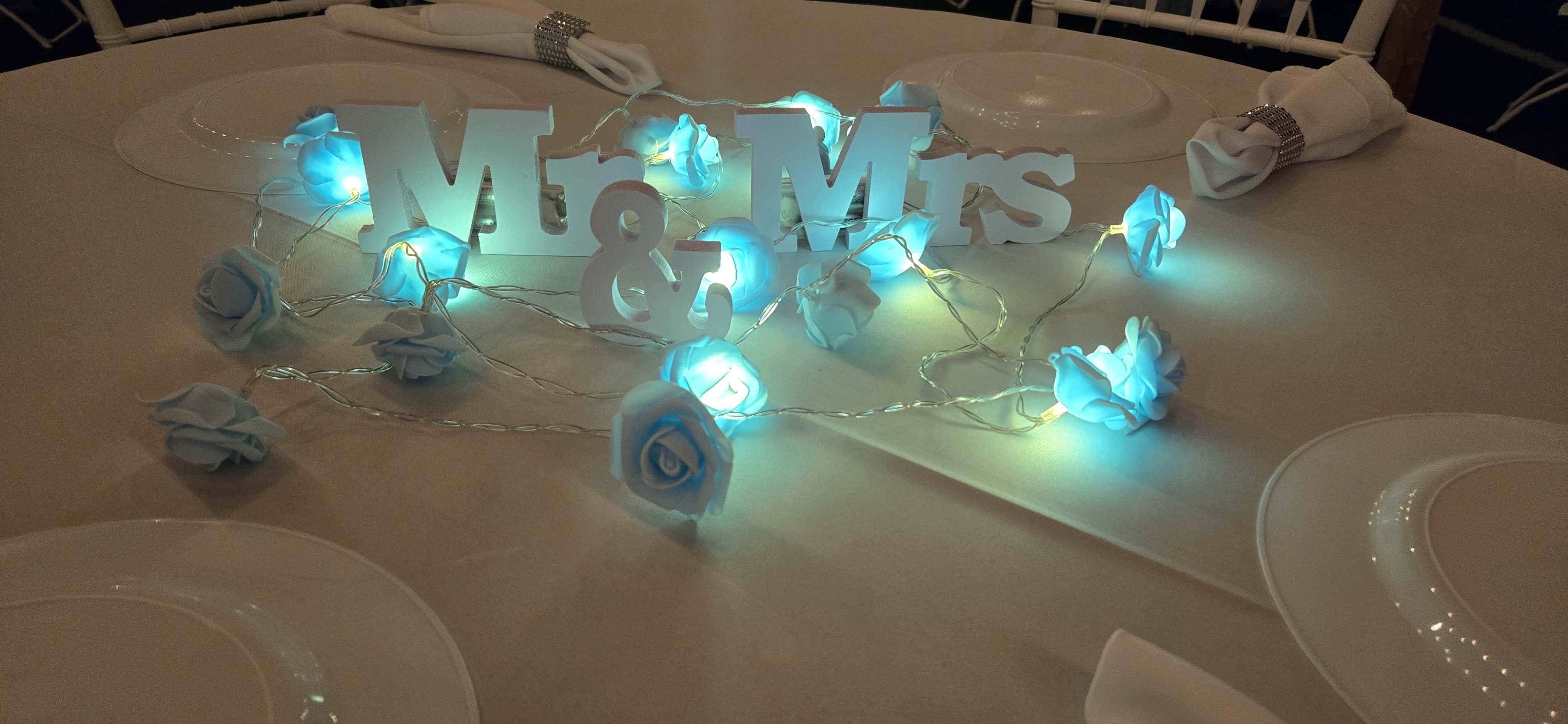 A table setting featuring a "Mr & Mrs" decoration surrounded by glowing blue rose decorations and soft white lights.