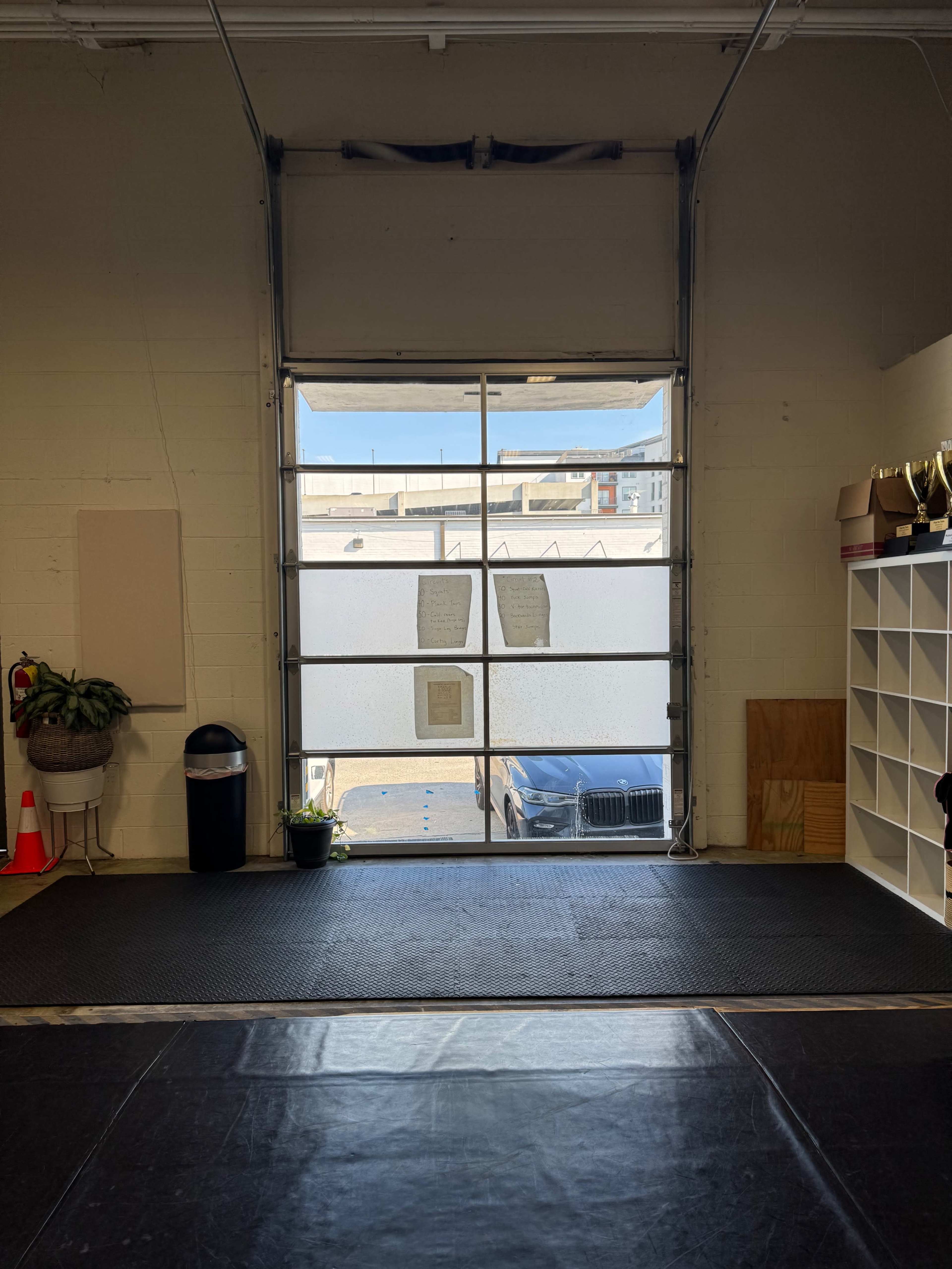 The image shows an indoor space with a large garage door partially open, revealing a view of a parked vehicle and outdoor area beyond.