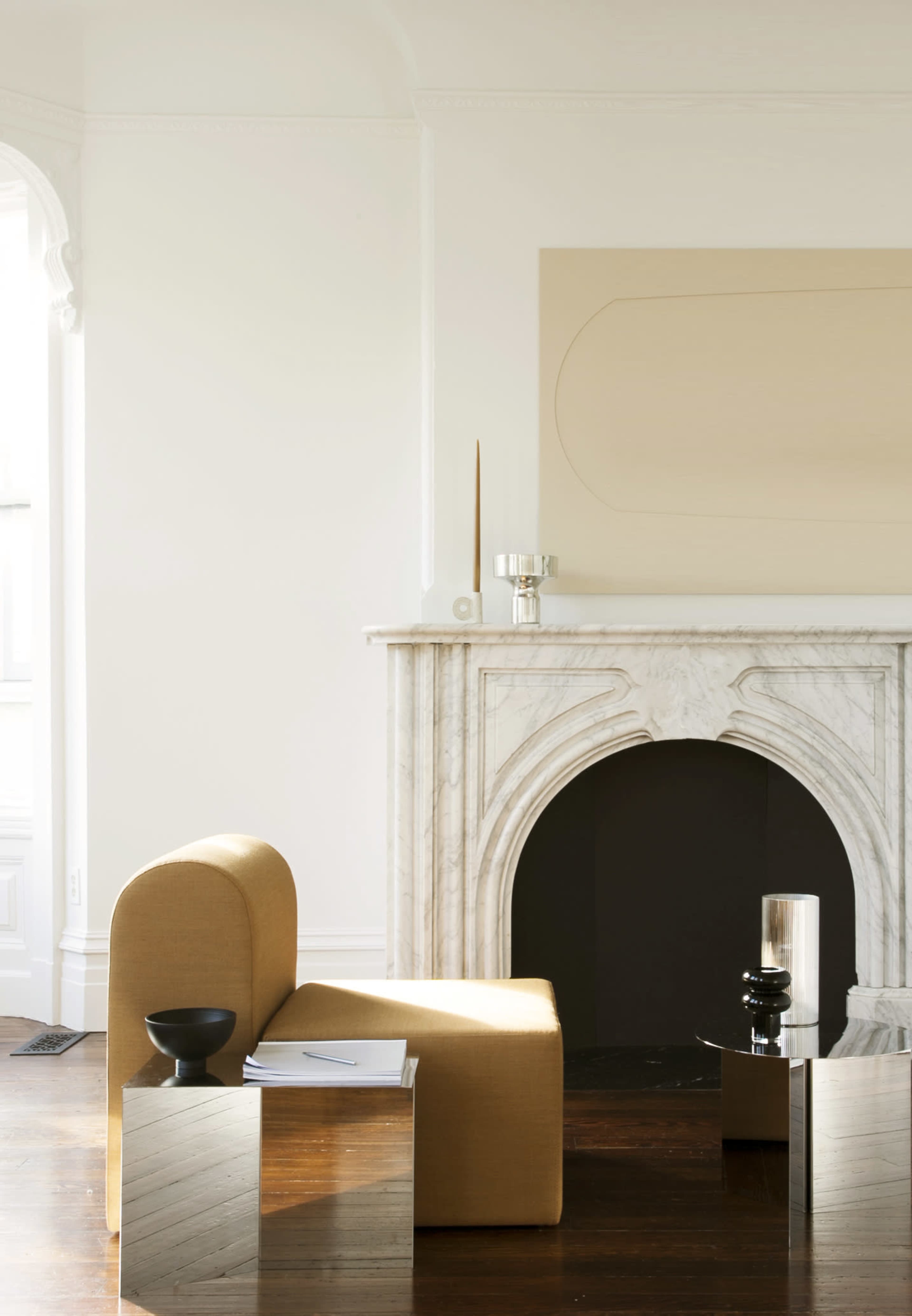 A minimalistic living room features a beige curved chair next to a marble fireplace, with a dark decorative object on a glass table.