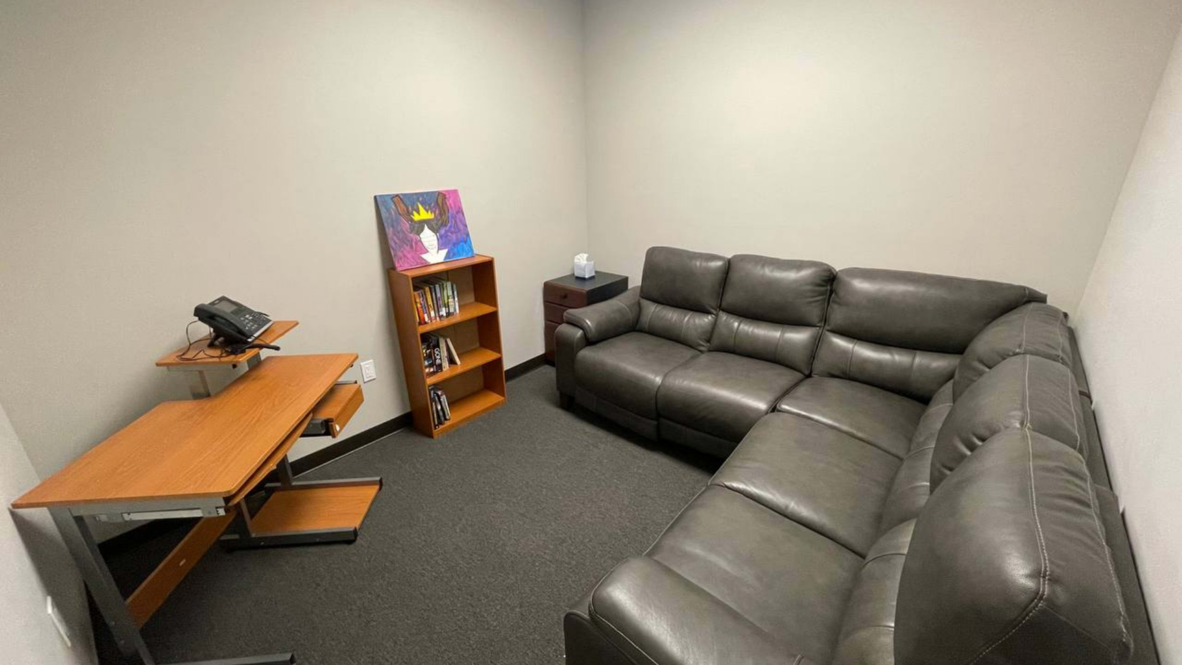 The image shows a small, private room furnished with a dark sectional sofa, a wooden desk with a telephone, and a bookshelf containing several books and a colorful painting on the wall.