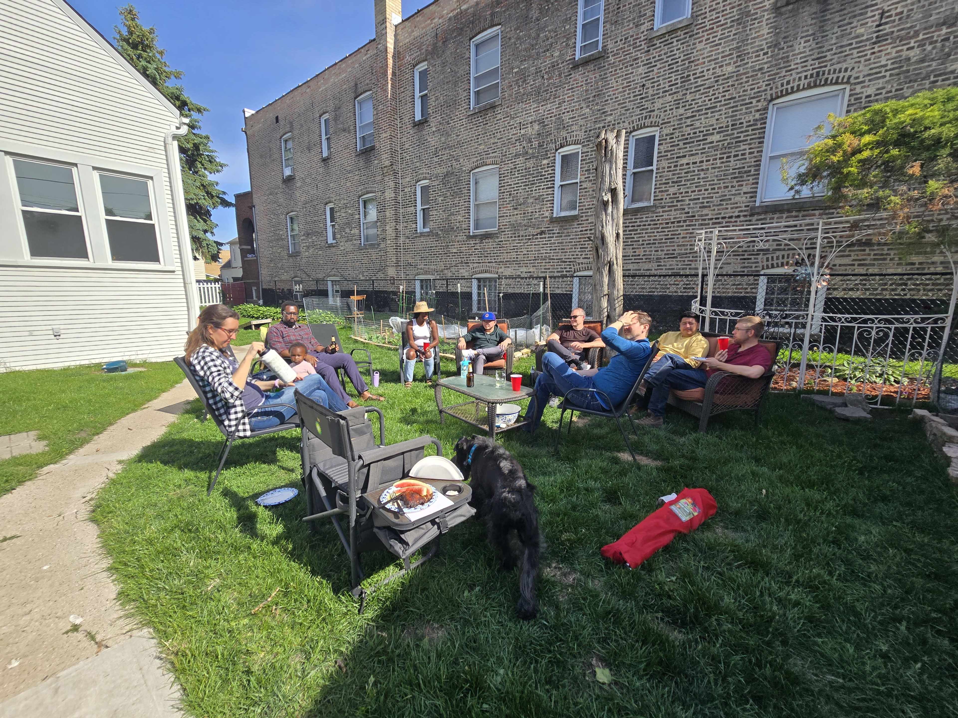 A group of people are sitting in a grassy area outside a building, engaged in conversation and socializing on a sunny day.