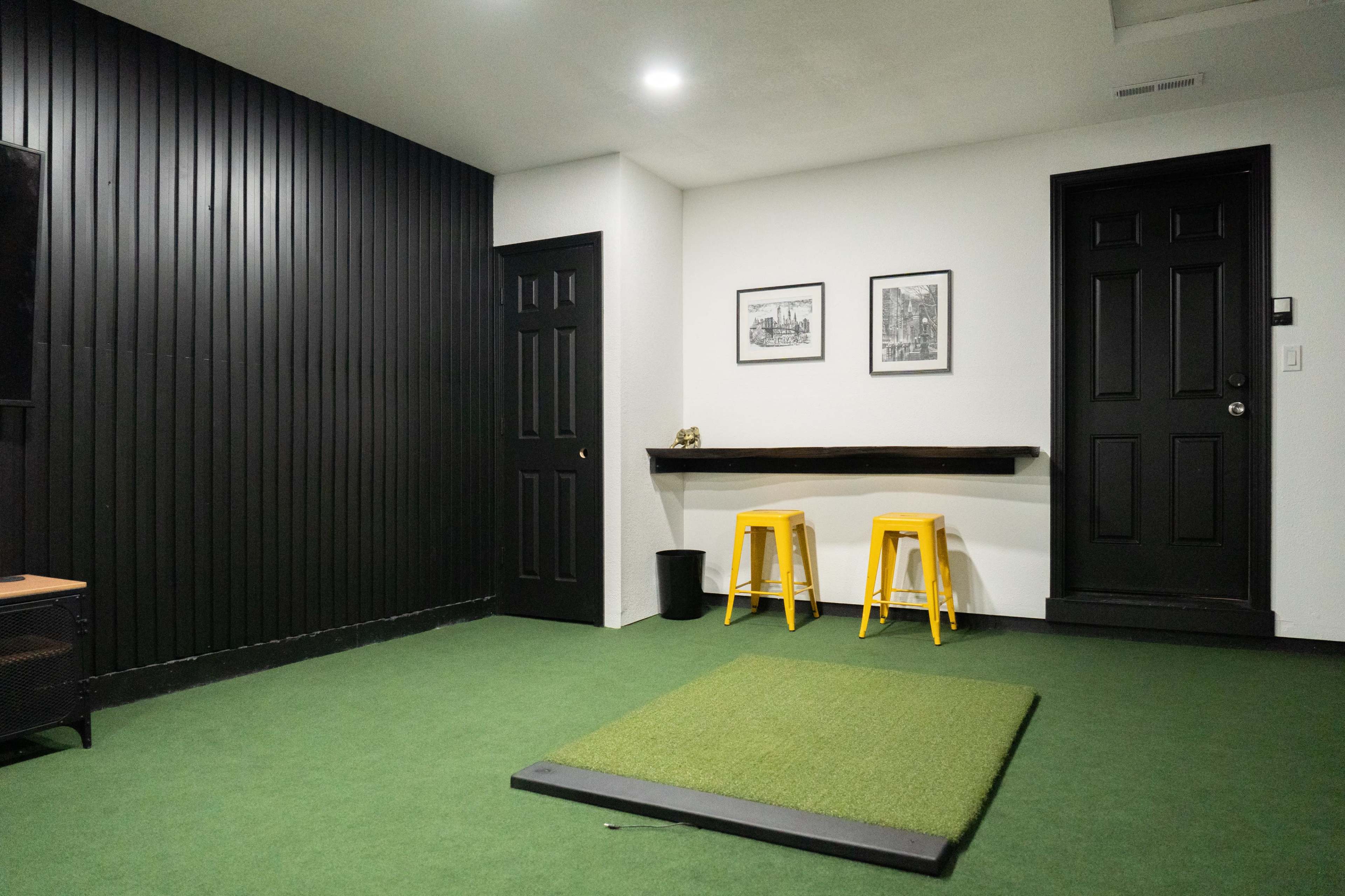 The room features a green carpeted area with a small, raised platform, two yellow stools, a wall-mounted shelf, and black doors against a dark wood-paneled wall.