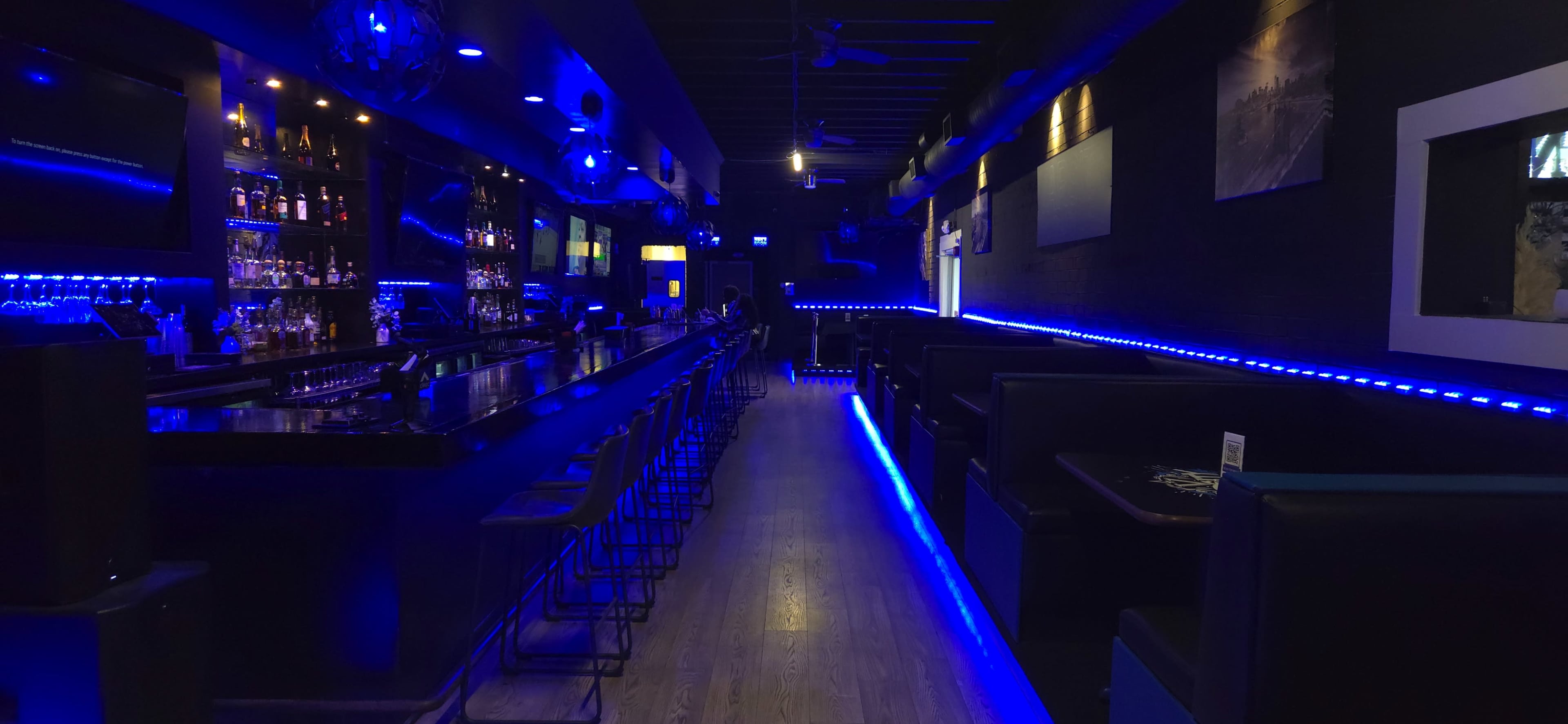 A dimly lit bar features a long counter with high stools, illuminated by blue LED lights lining the walls and bar area.