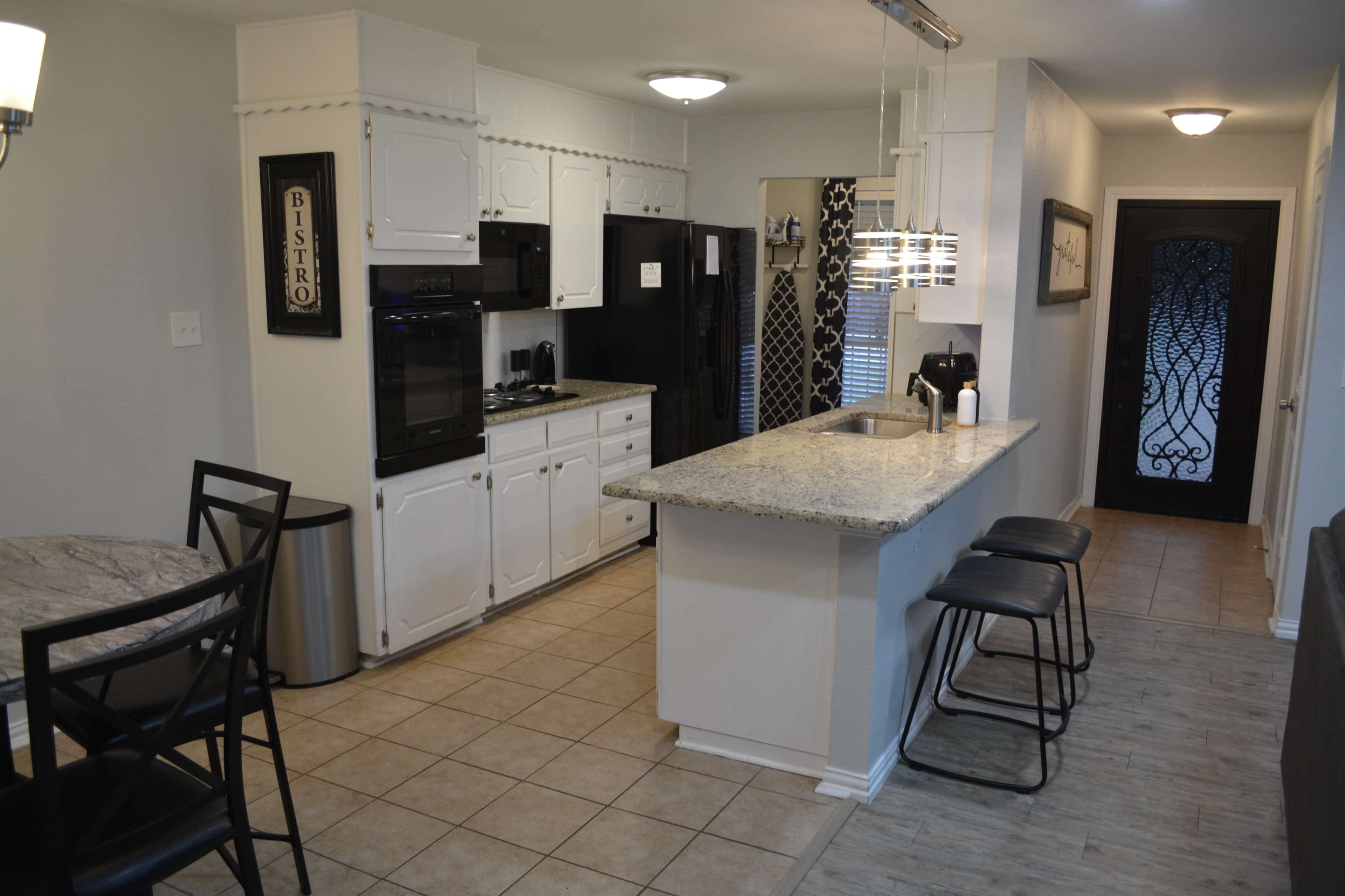 The image shows a modern kitchen with white cabinetry, granite countertops, and a black refrigerator, adjacent to a dining area with a small round table and black bar stools.