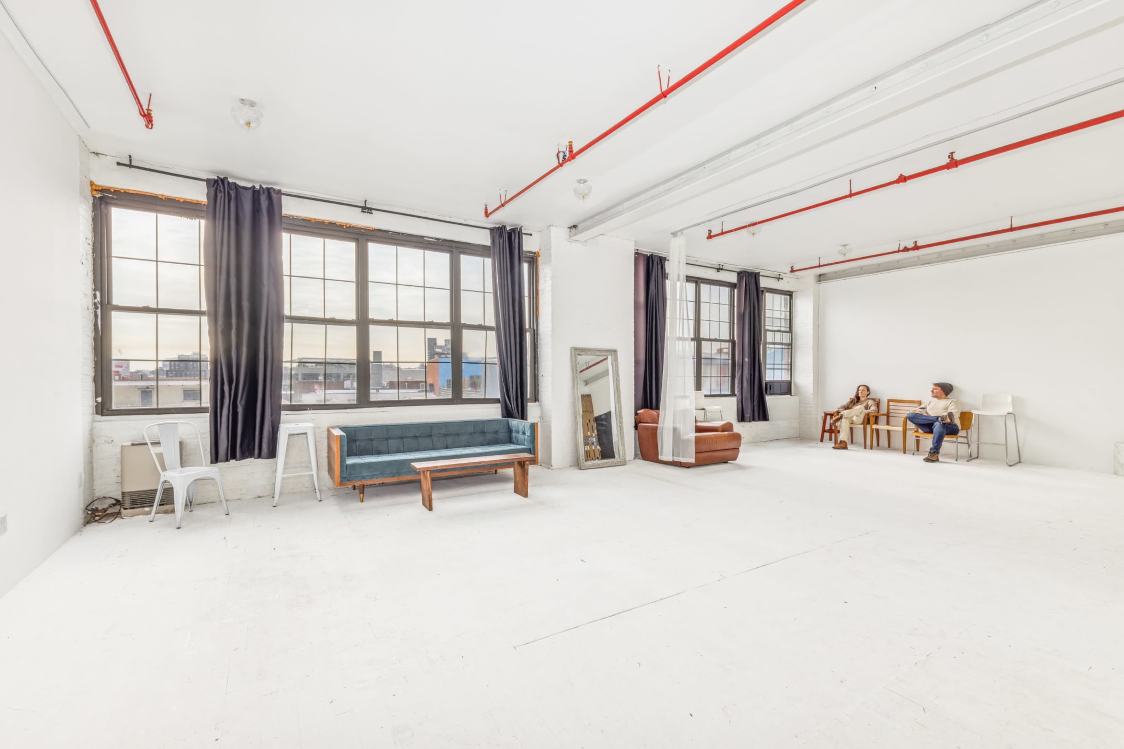 A spacious, brightly lit studio features large windows, minimalist furniture, and a white floor.