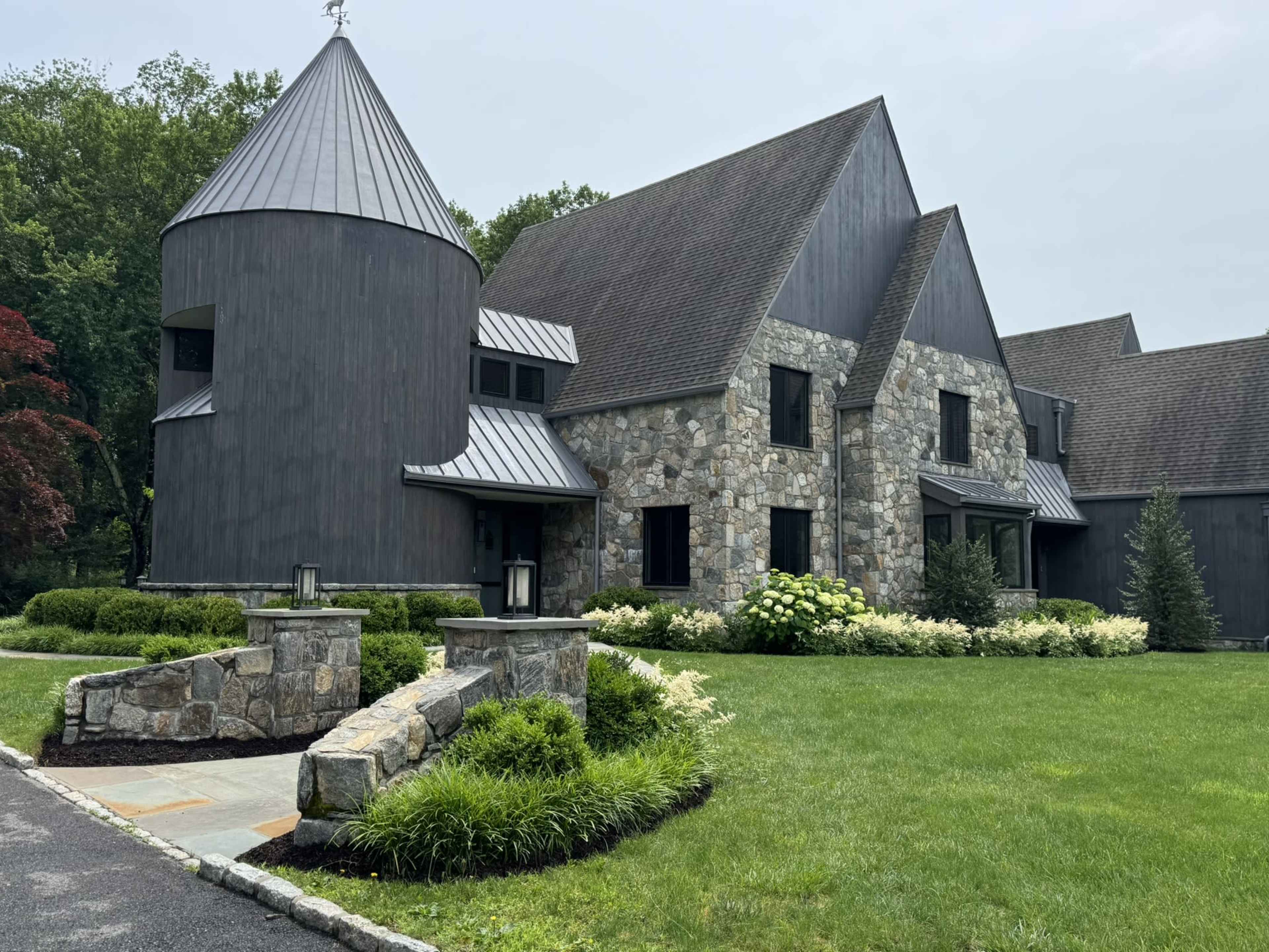 The image shows a modern house featuring a combination of stone and dark wood exteriors, with a unique turret-style structure and landscaped yard.