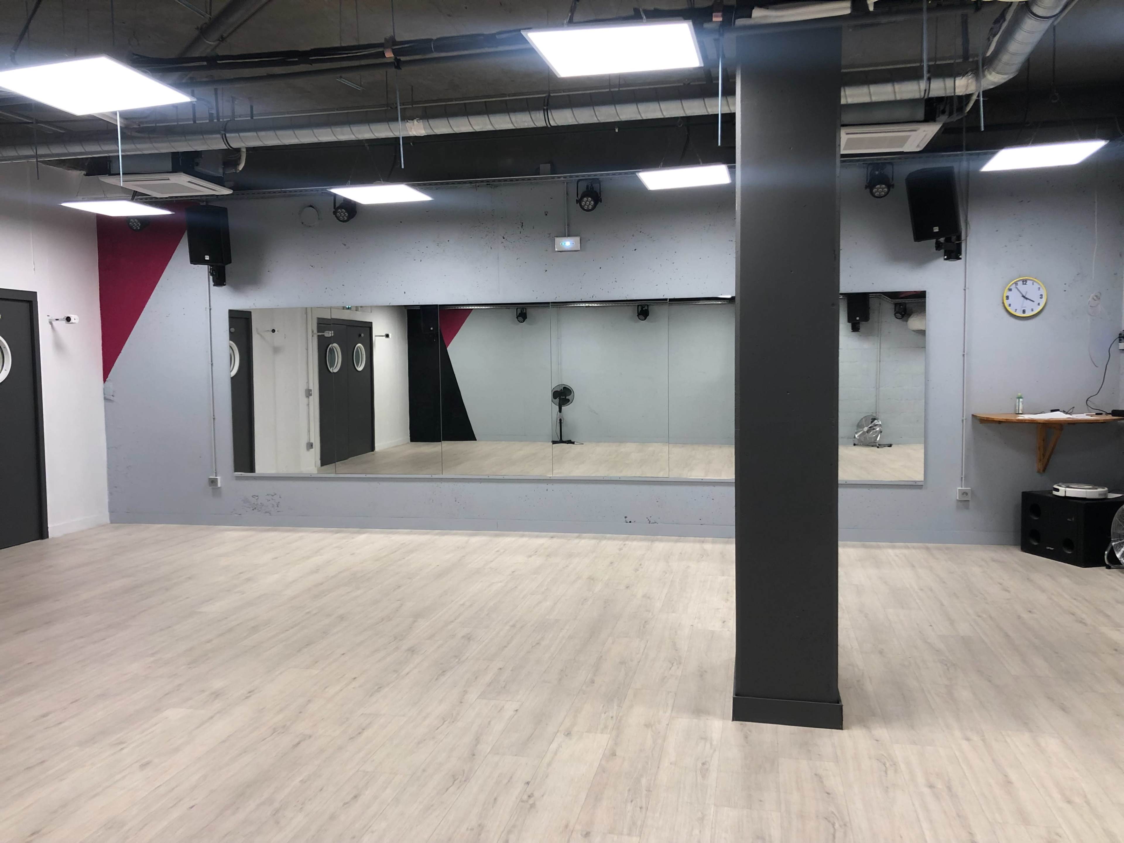 A spacious, empty studio with wooden flooring, mirrored walls, and a clock on one wall.