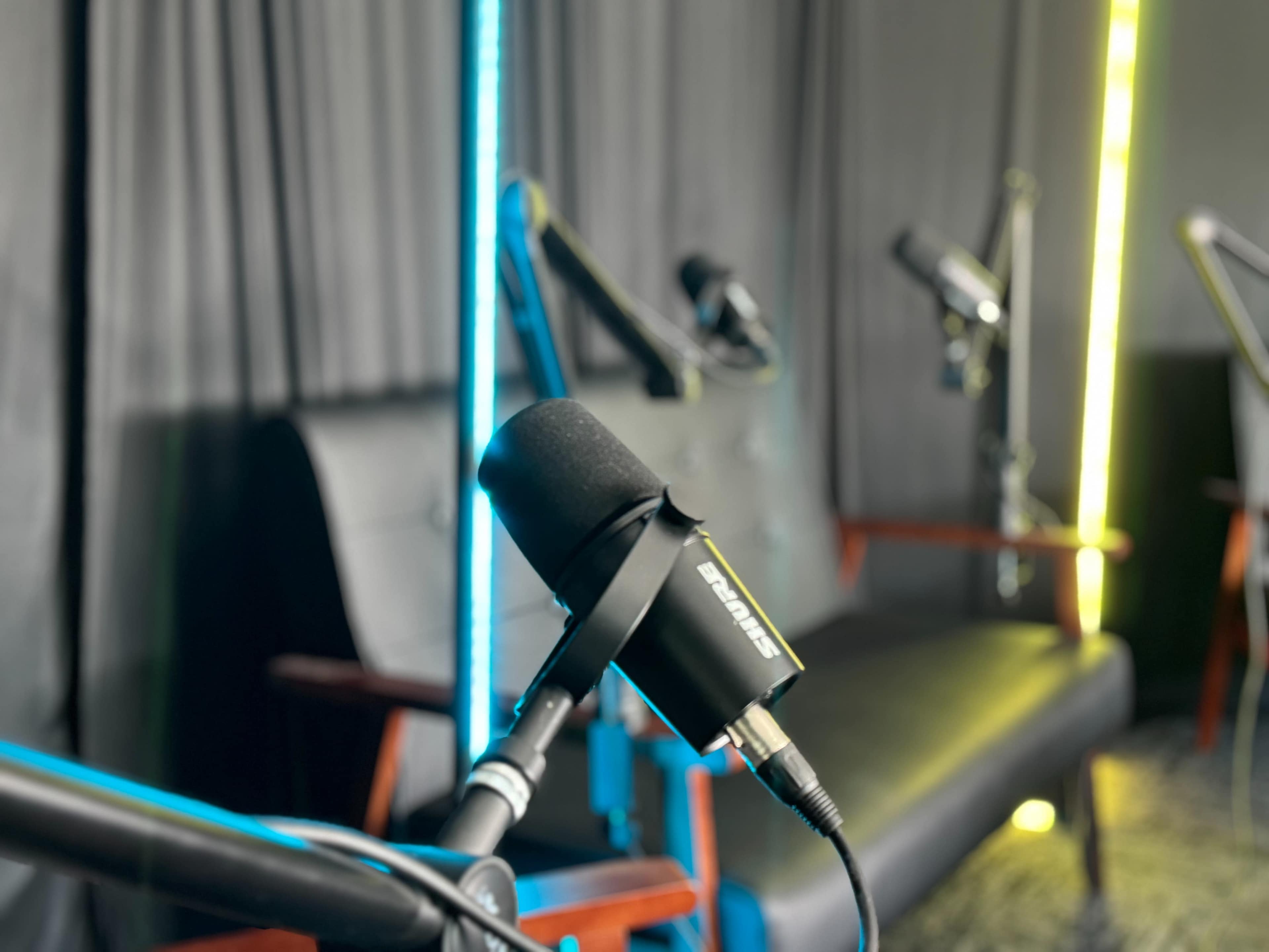 A close-up view of a microphone on a stand in a recording studio, with brightly lit background elements and empty seating.