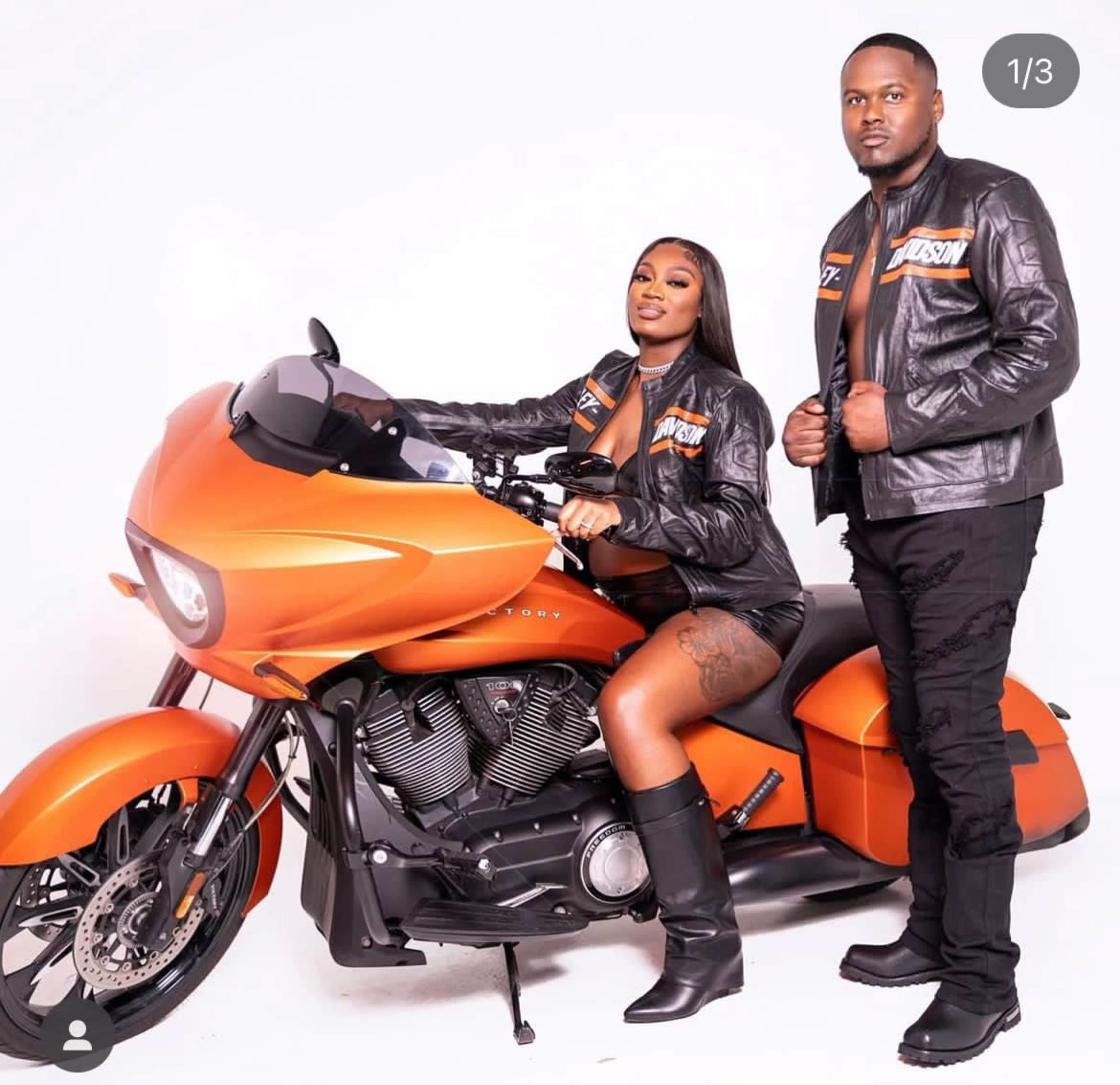A woman sitting on an orange motorcycle poses alongside a man in matching leather jackets.