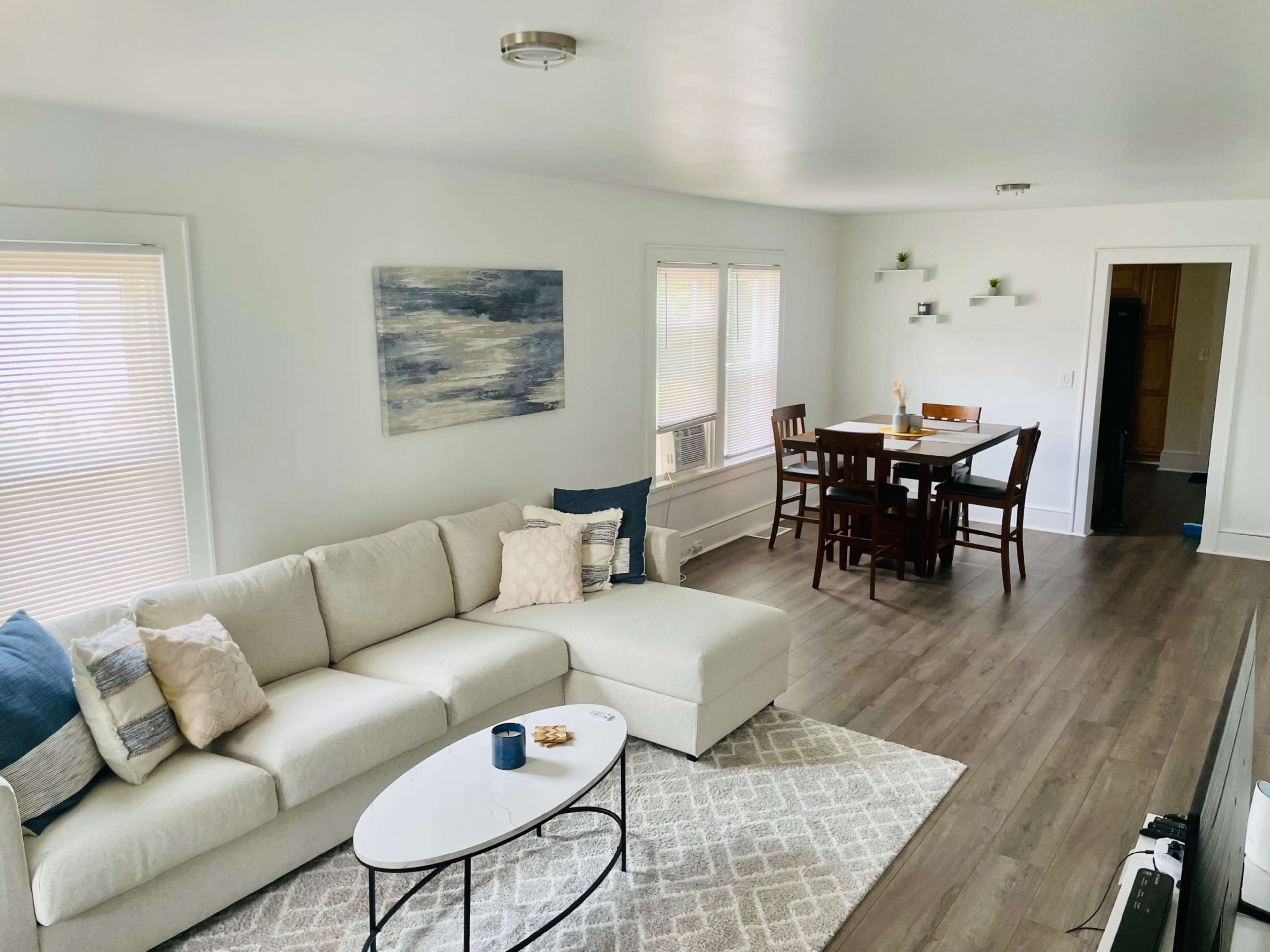 The image shows a bright living room with a light-colored sectional sofa, a coffee table, and a dining area with a wooden table and chairs.