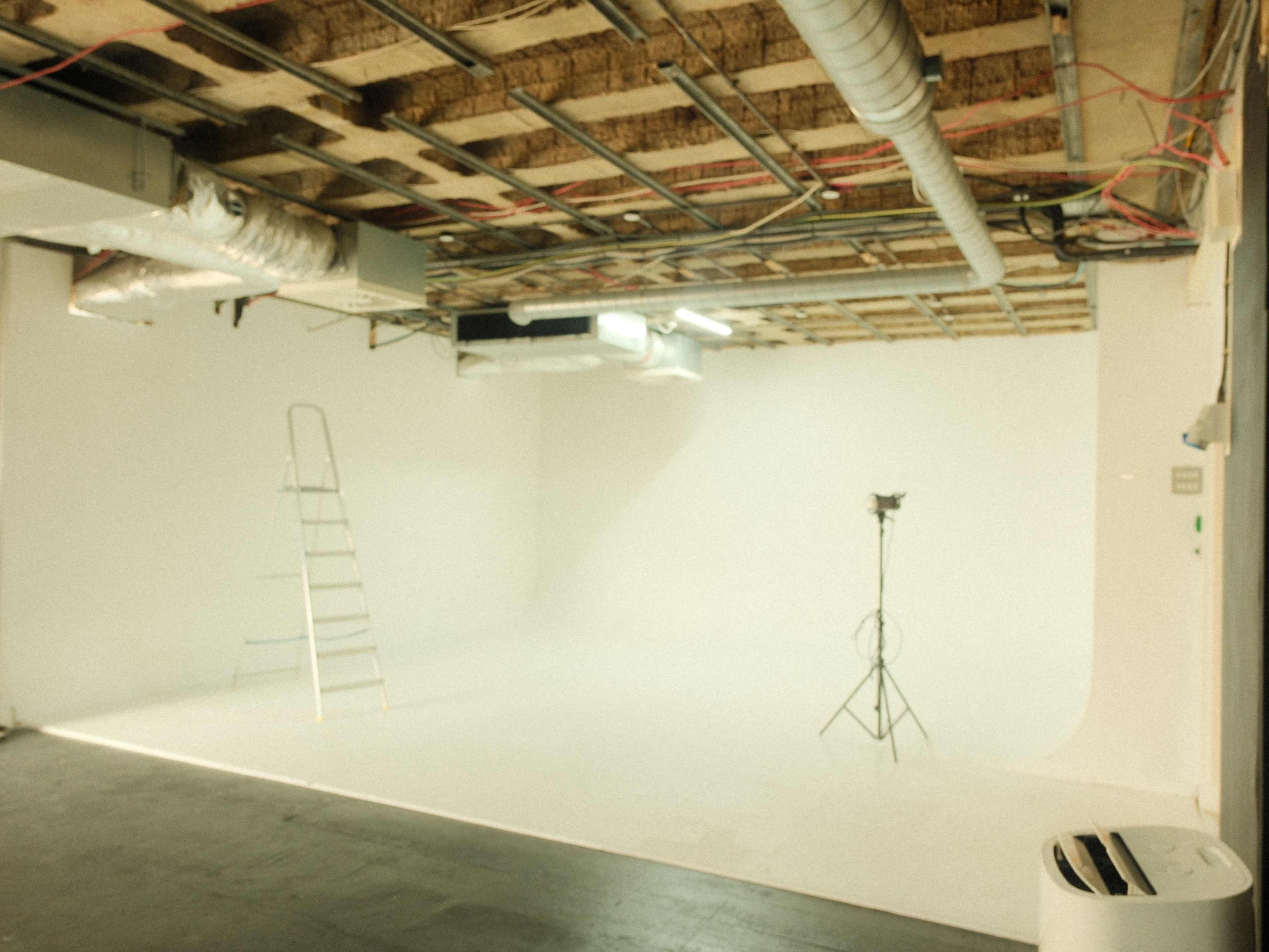 The image shows a spacious studio with a white backdrop, a metal ladder, and a lighting stand.