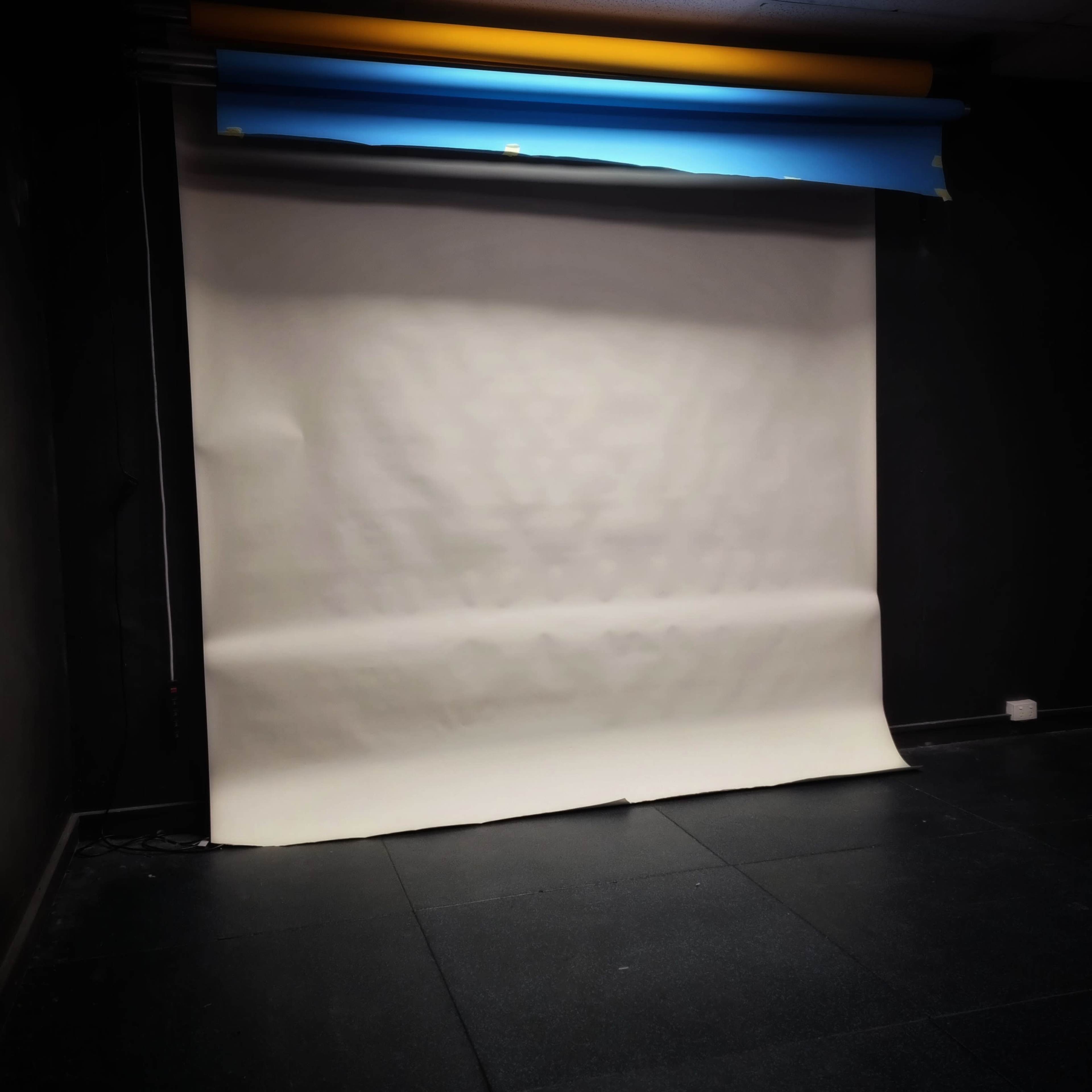 A blank photography backdrop made of white paper, set against a dark wall with some colored rolls of paper above it.