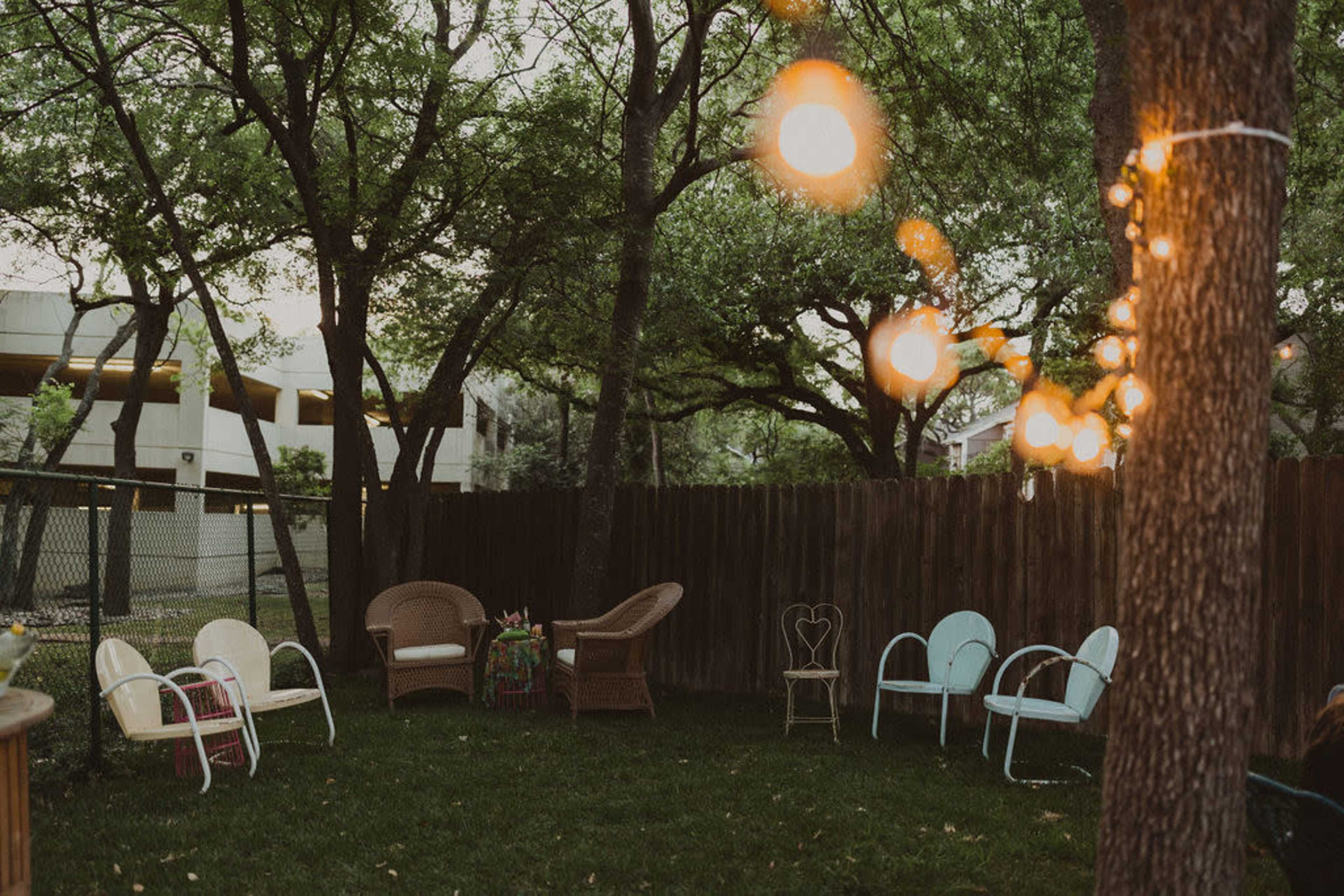 A backyard with several chairs arranged in a circle, illuminated by string lights, surrounded by trees and a wooden fence.
