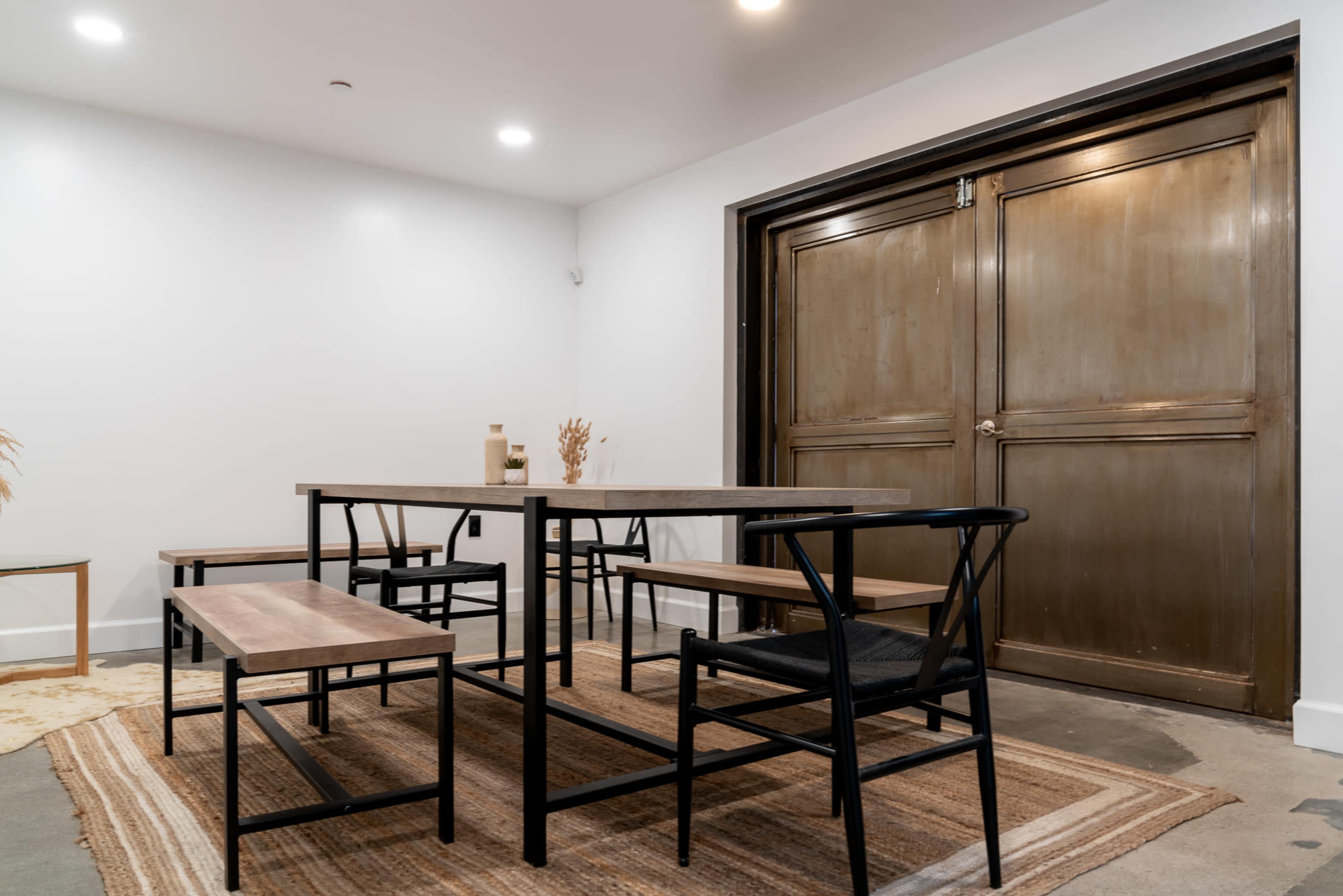 A dining area features a simple wooden table with benches, set on a woven rug, adjacent to a large closed door.