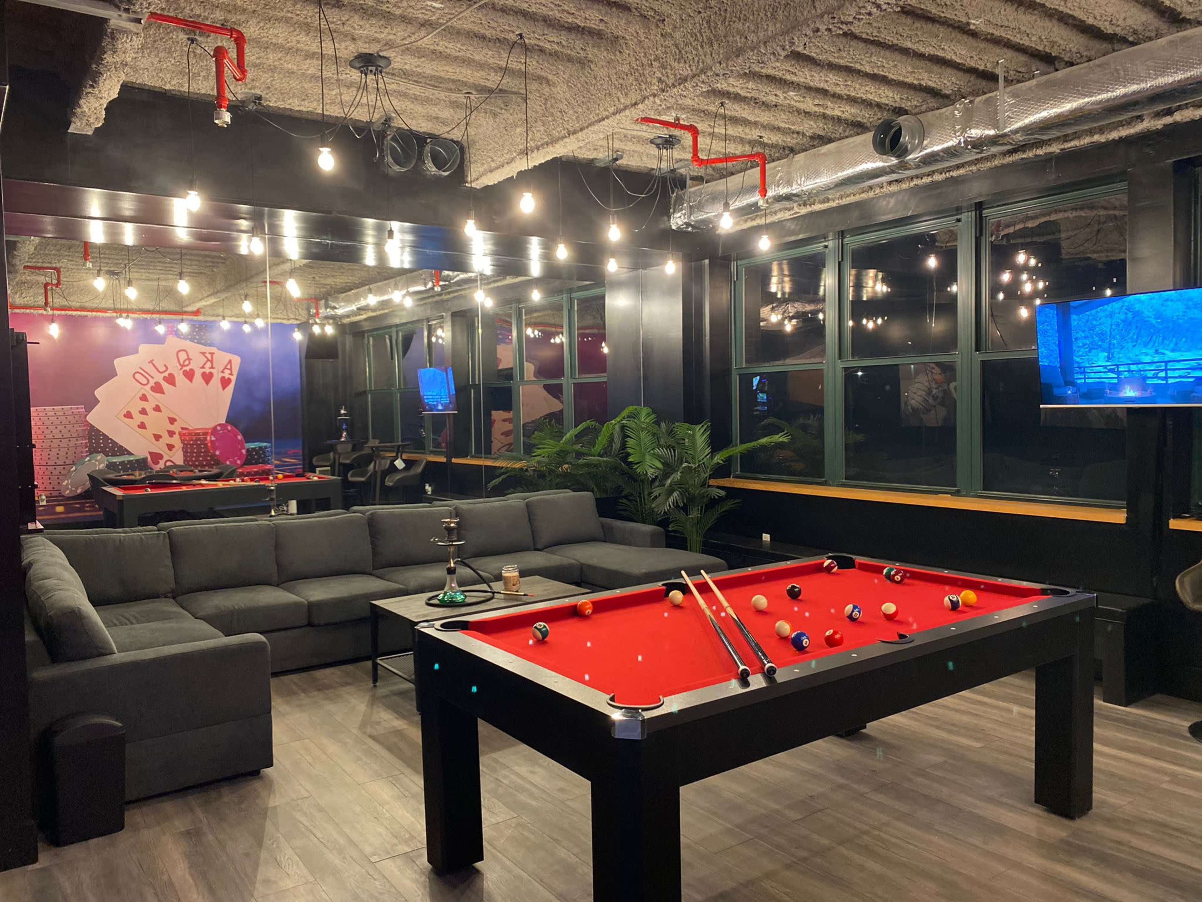 A game room featuring a red pool table, a large gray sectional sofa, and wall-mounted televisions, all illuminated by pendant lights.