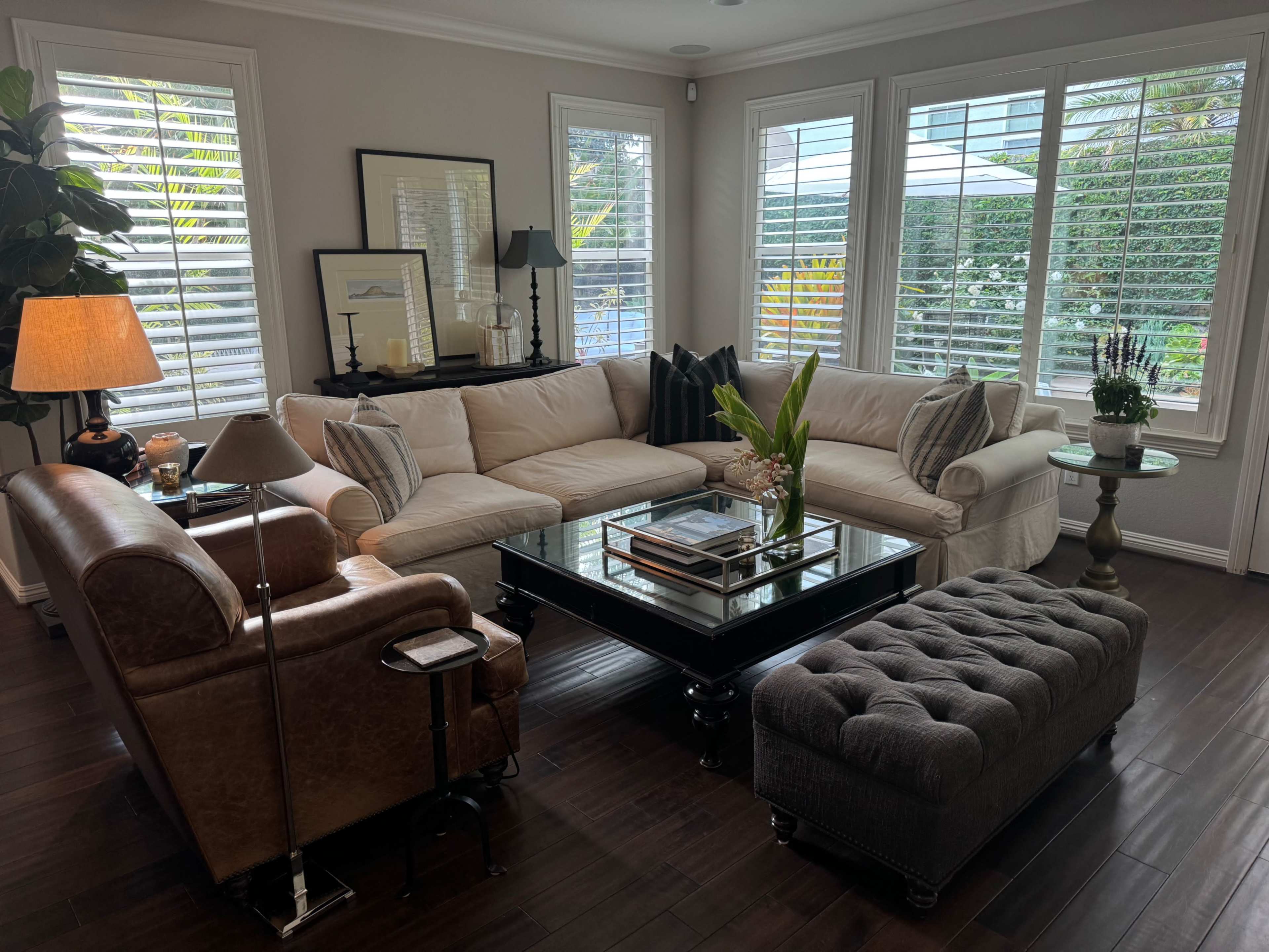 A bright living room features a sectional sofa, a coffee table, and a combination of plants and decorative elements near large windows with shutters.