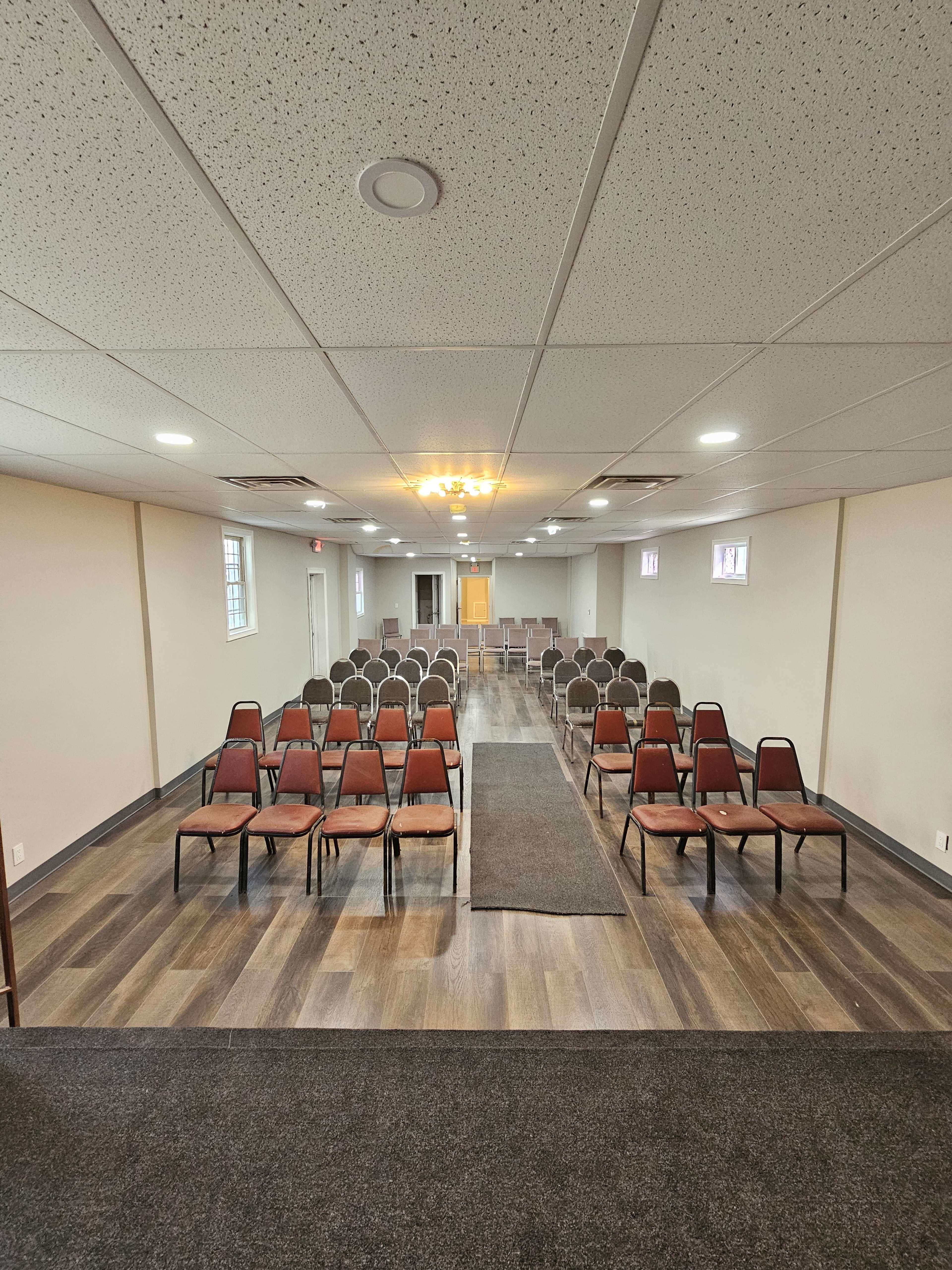 The image shows a meeting room set up with rows of chairs facing a central aisle, with walls painted in neutral tones and a carpeted floor.