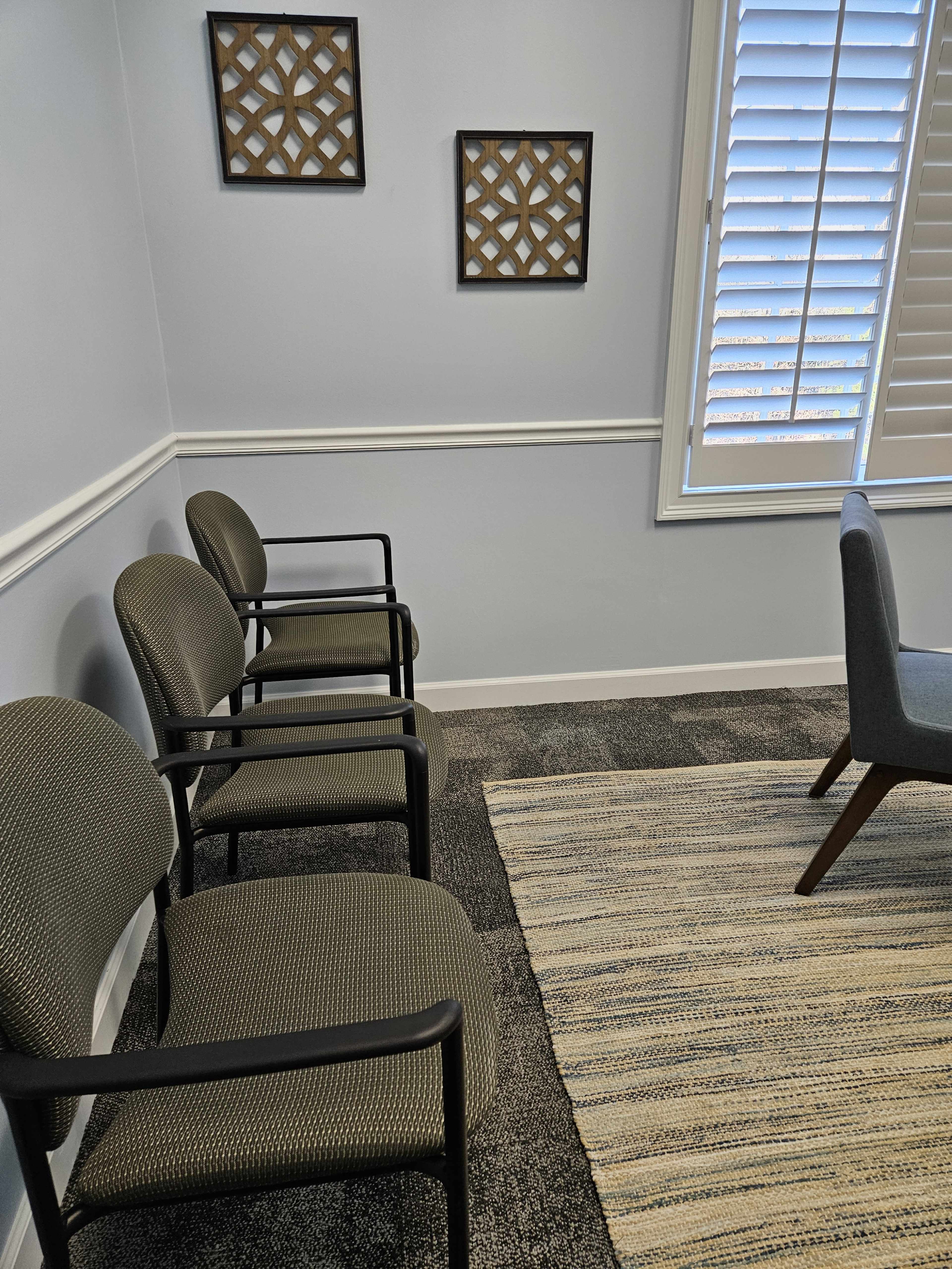 The image shows a small room with three chairs along one wall, a patterned rug on the floor, and two framed decorations on another wall beside a window with shutters.