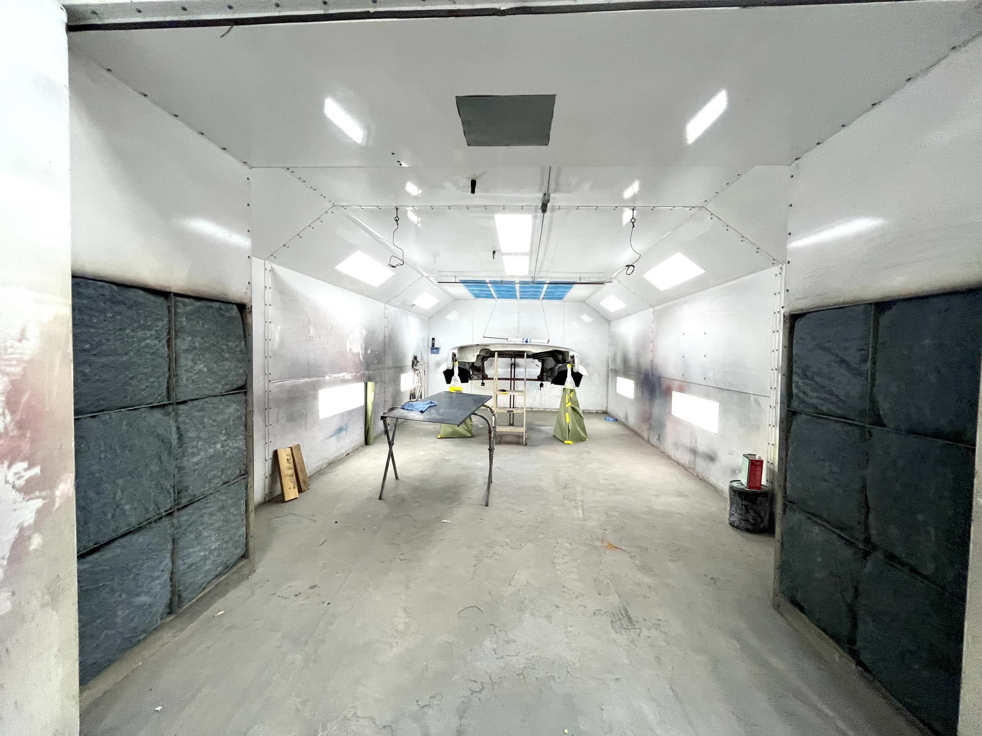 The image shows an empty paint booth with large sliding doors, a spray setup in the center, and bright overhead lights.