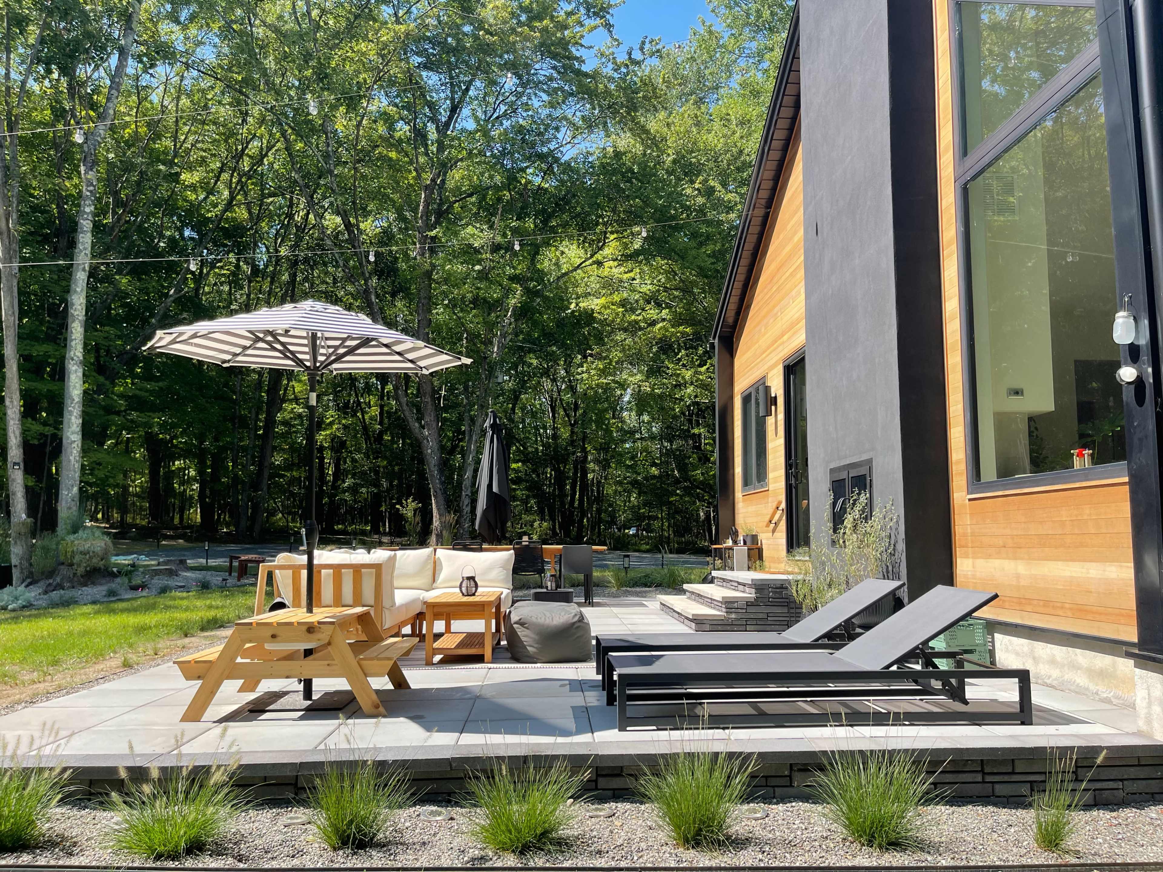 A patio area features a wooden lounge set, an umbrella, and two chaise loungers surrounded by trees.