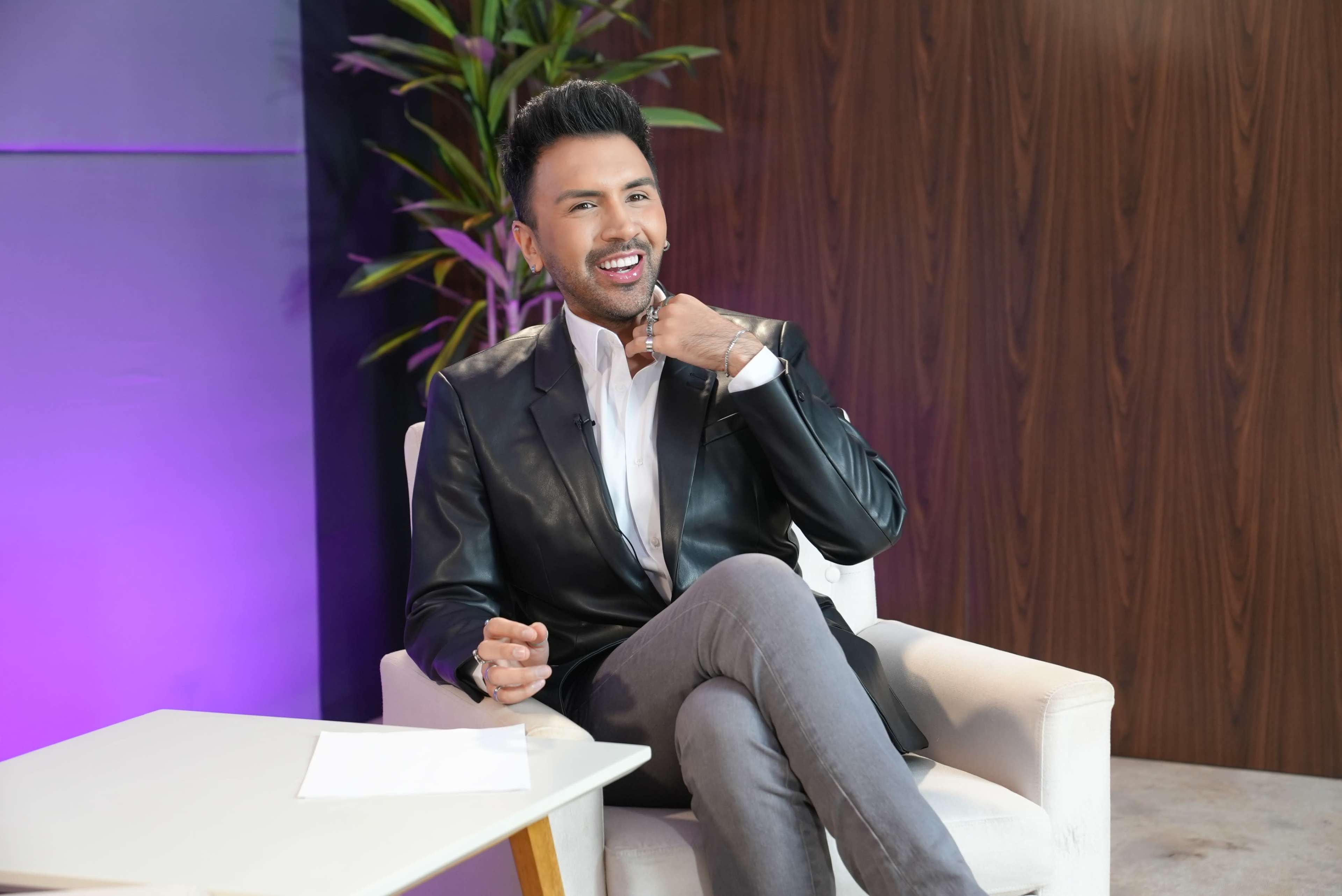 A man sits on a chair, smiling and gesturing while wearing a black leather jacket and gray pants, with a plant and a wooden wall in the background.