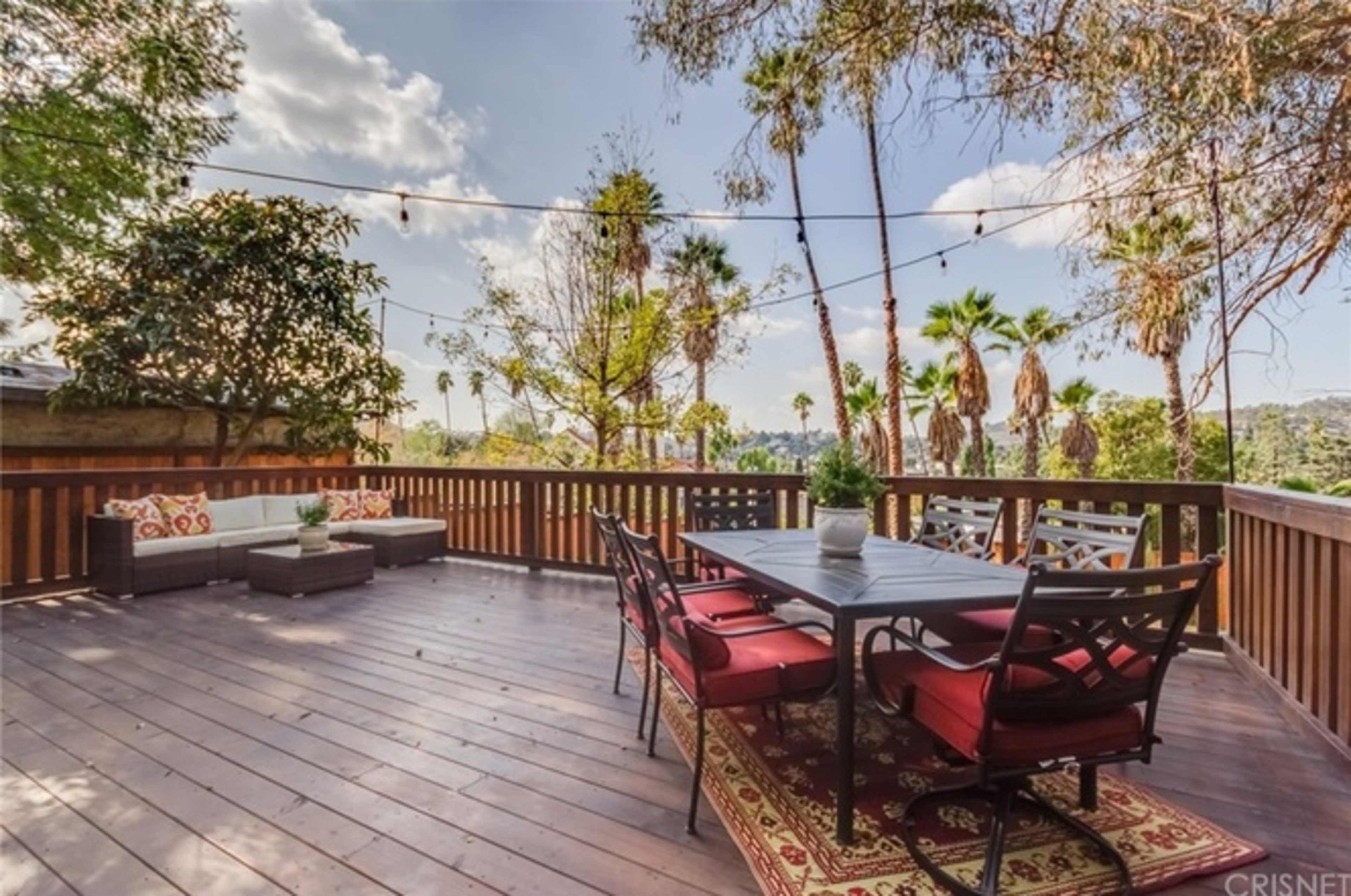 A wooden deck features a dining table with black chairs and a potted plant, surrounded by palm trees and a mountainous backdrop under a partly cloudy sky.