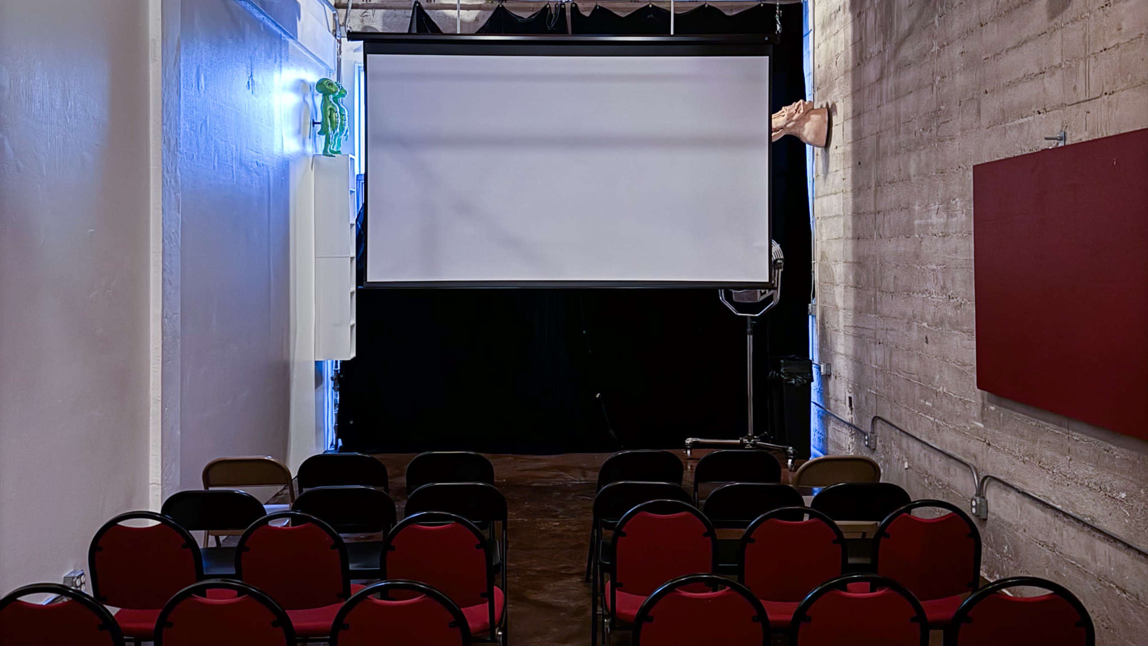 A small auditorium with red chairs arranged in rows facing a large blank projection screen, set against a dark backdrop.