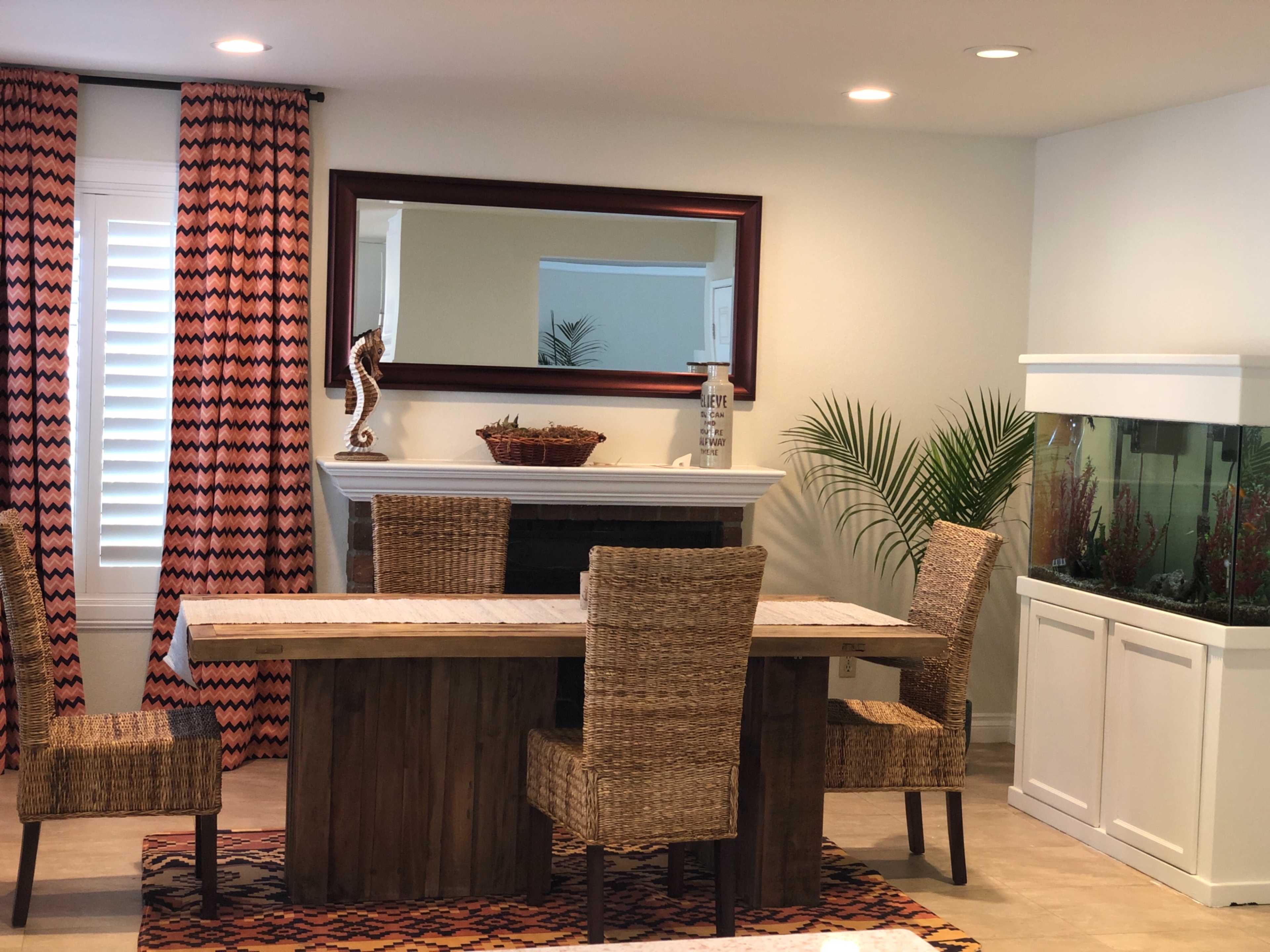 A dining area features a wooden table surrounded by wicker chairs, with a mirror, decorative elements, and an aquarium in the background.