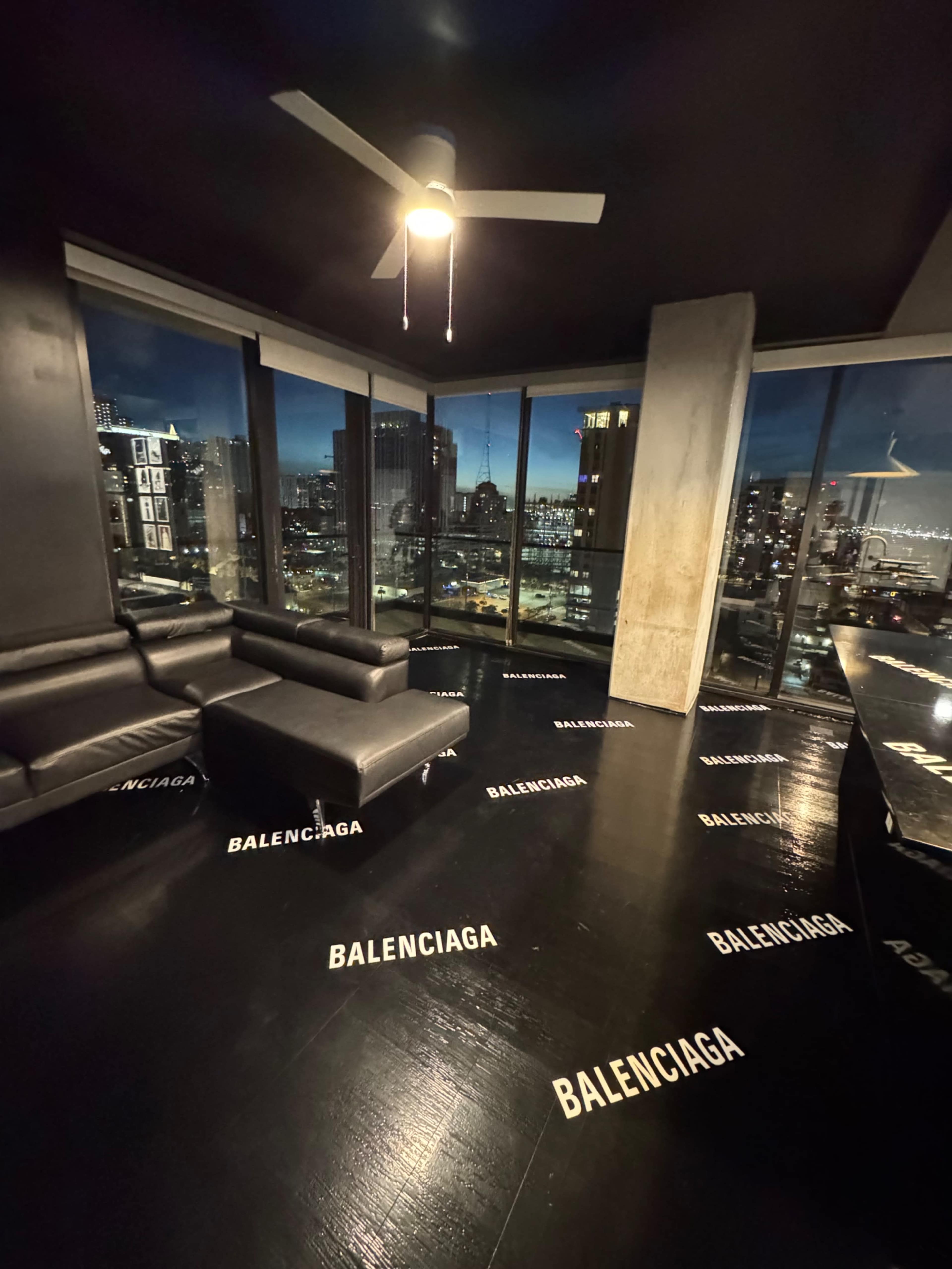 The image shows a modern living space with large windows overlooking a city skyline at dusk, featuring a black floor marked with the word "BALENCIAGA" and minimalist furniture.