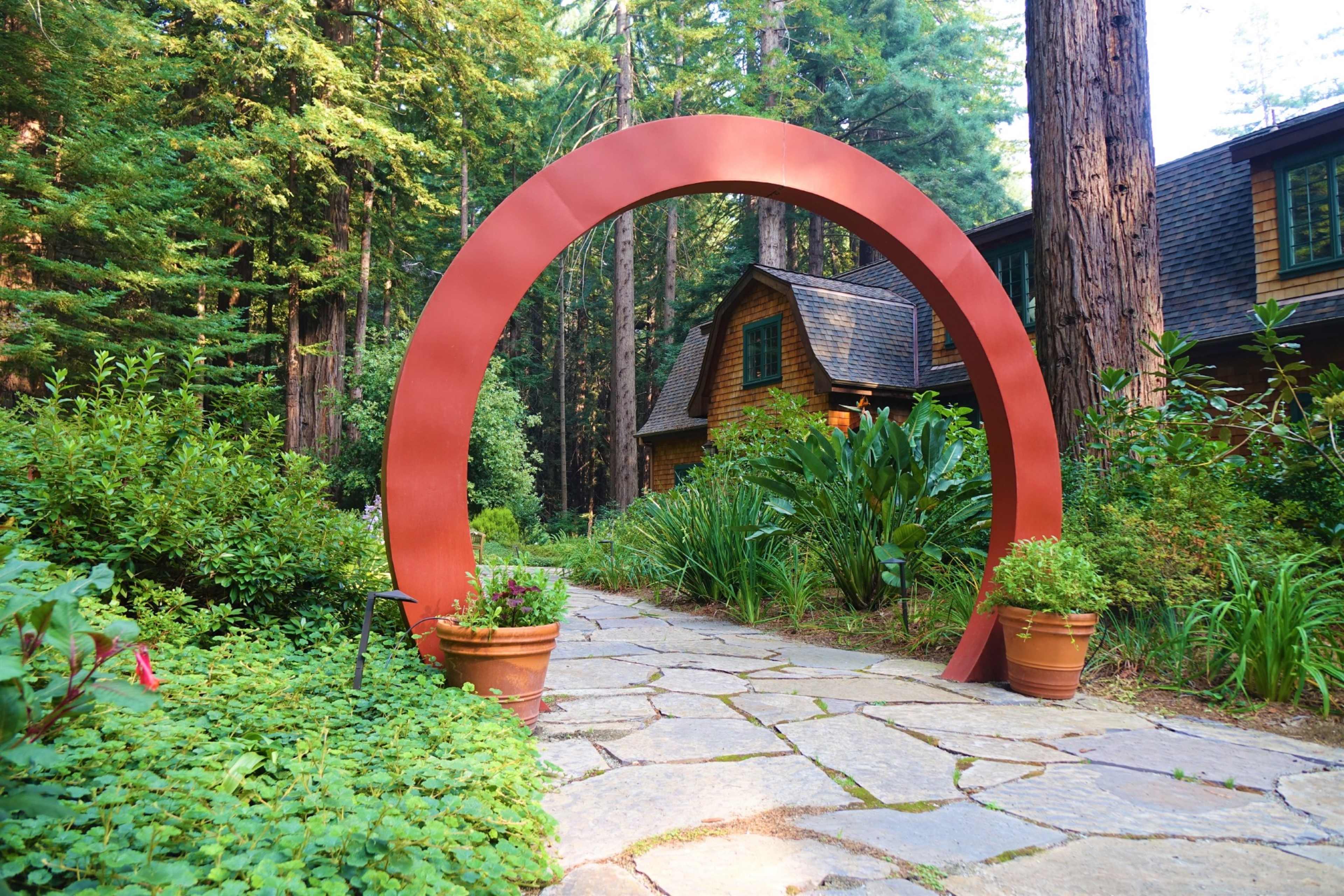 A red arch frames a stone pathway leading to a house nestled among tall trees and lush greenery.