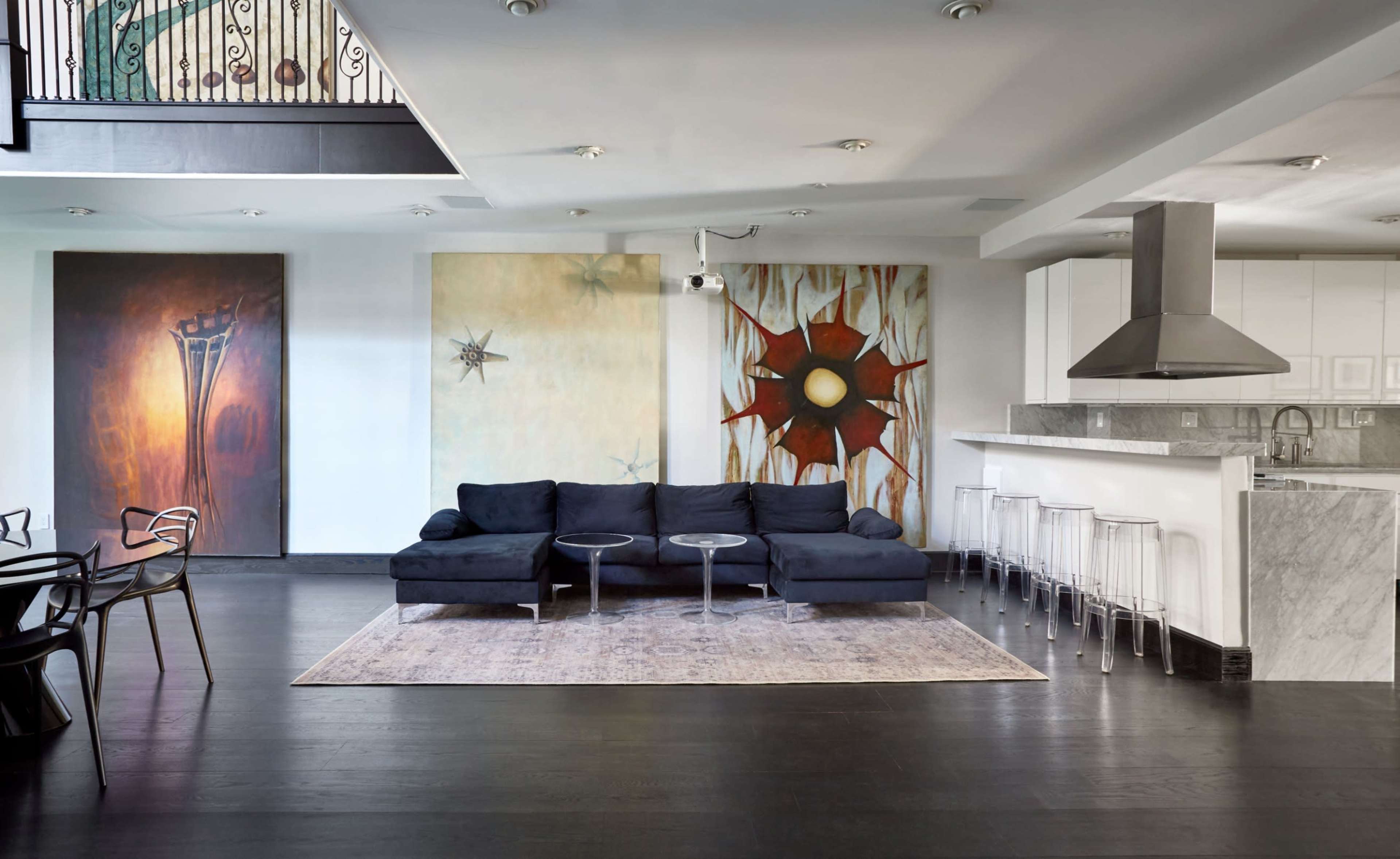 A modern living room features a dark sectional sofa, two round side tables, and a kitchen area with a marble counter and clear stools, complemented by large abstract paintings on the walls.