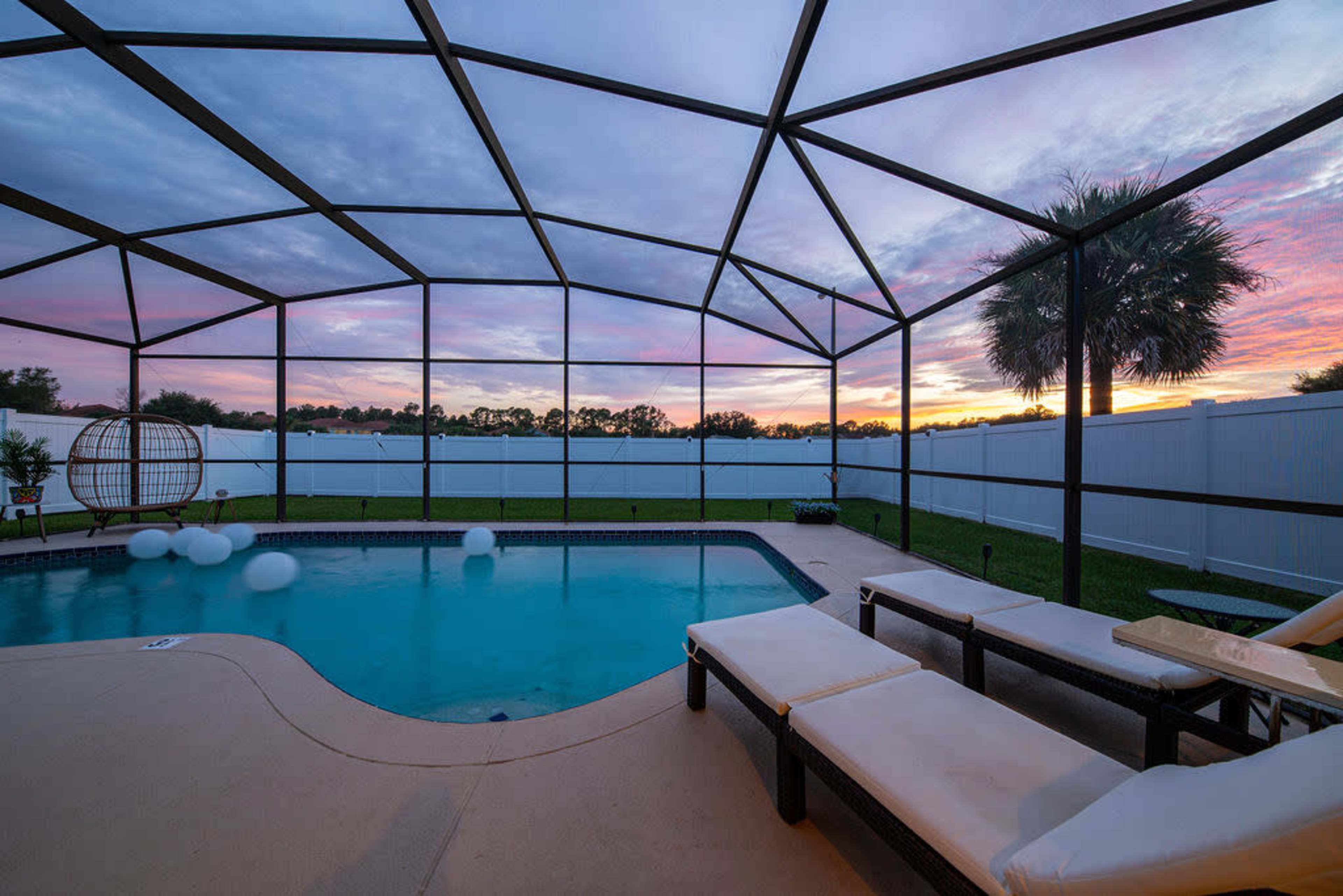 The image shows a swimming pool enclosed by a screen with lounge chairs beside it, under a colorful sunset sky.