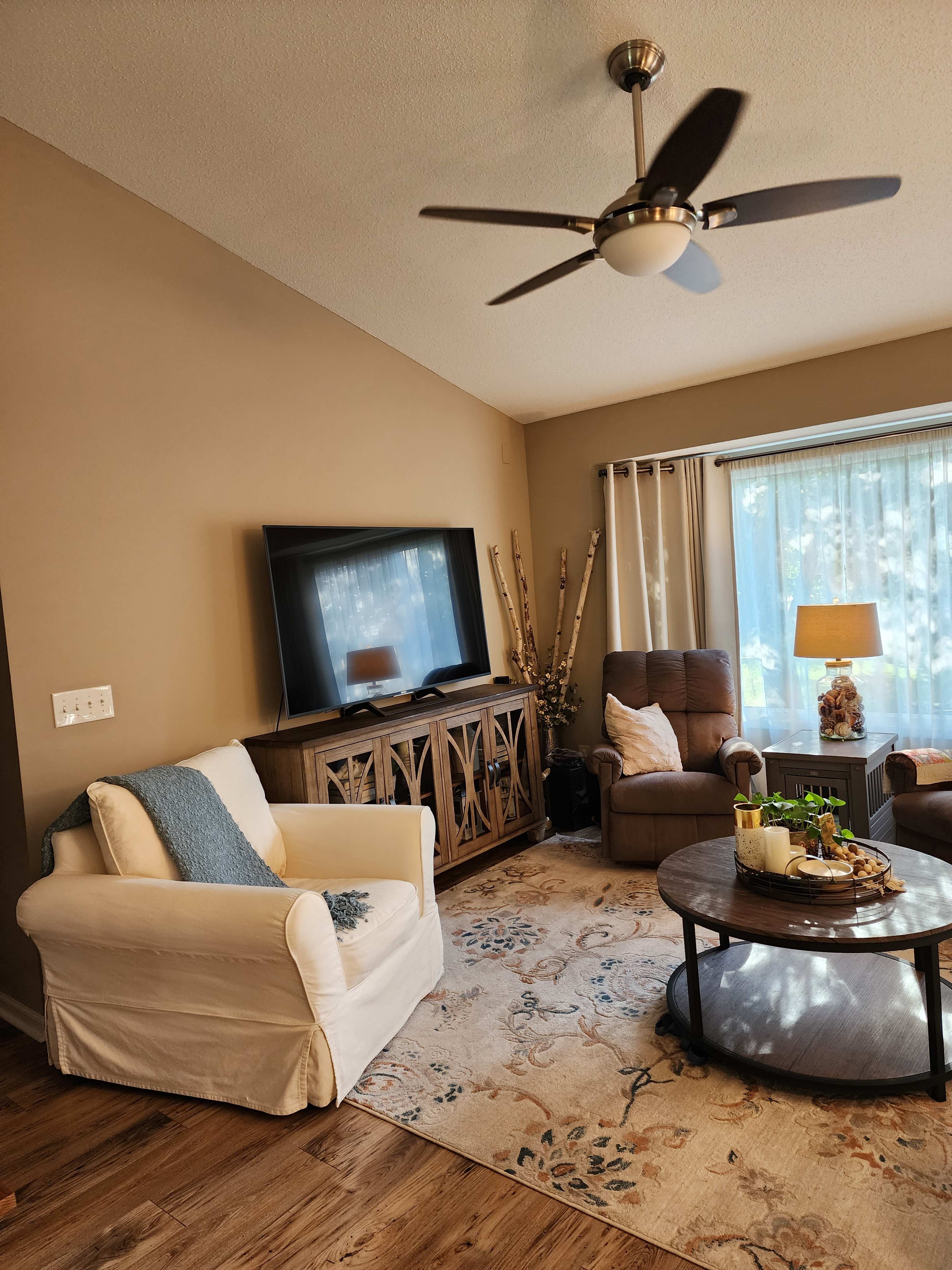 The image depicts a cozy living room featuring a television mounted on the wall, a circular coffee table in the center, and a beige sofa with a blue throw pillow.